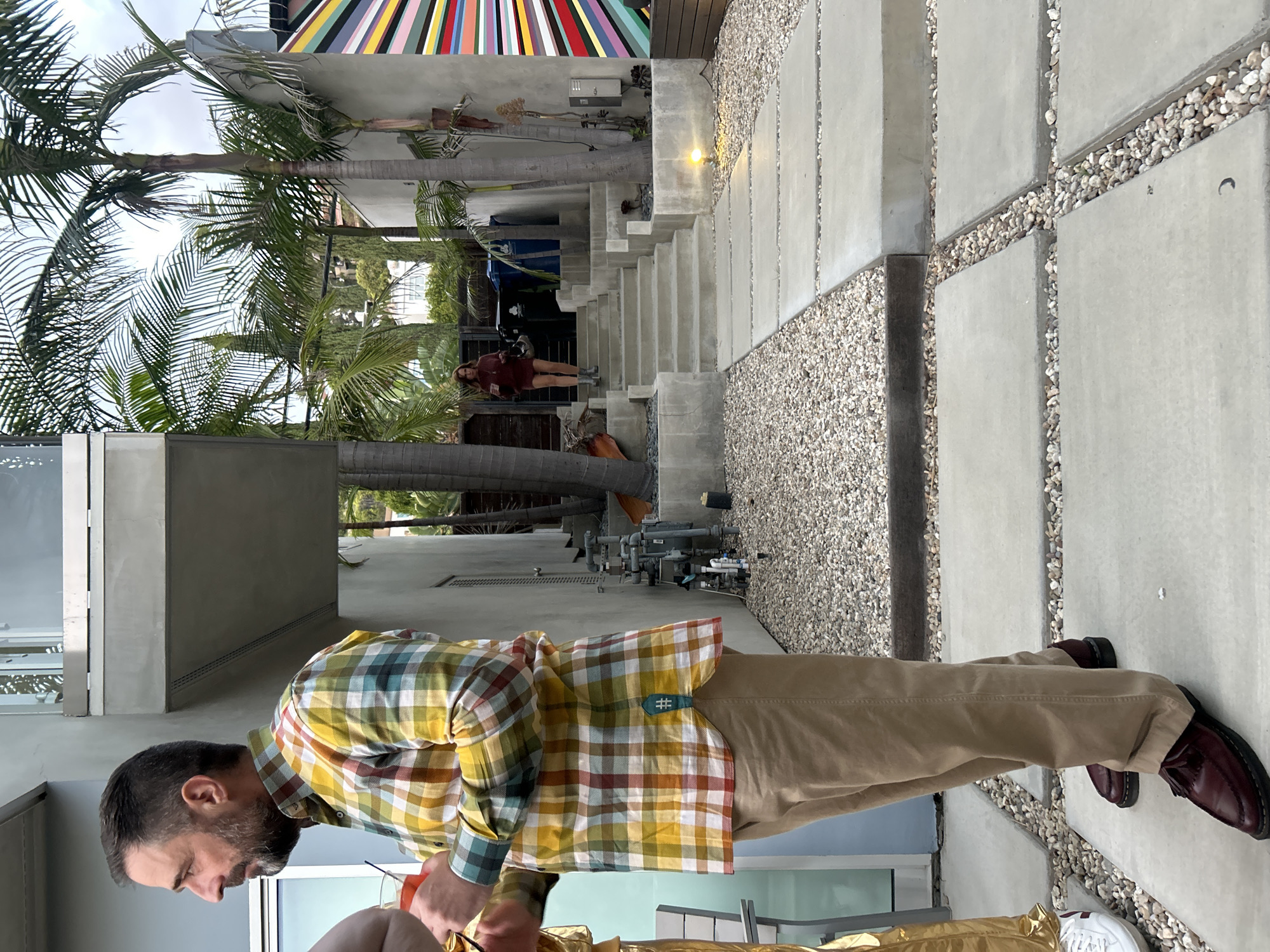 The image shows a man with a beard wearing a plaid shirt and khakis standing near a building. The background contains palm trees, a walkway made of concrete slabs and pebbles, stairs leading up to another part of the building, and another person standing on the stairs. The building in the background has a colorful striped design on the top. The location is likely West Hollywood, United States of America, based on the architectural style and vegetation typical of the area.