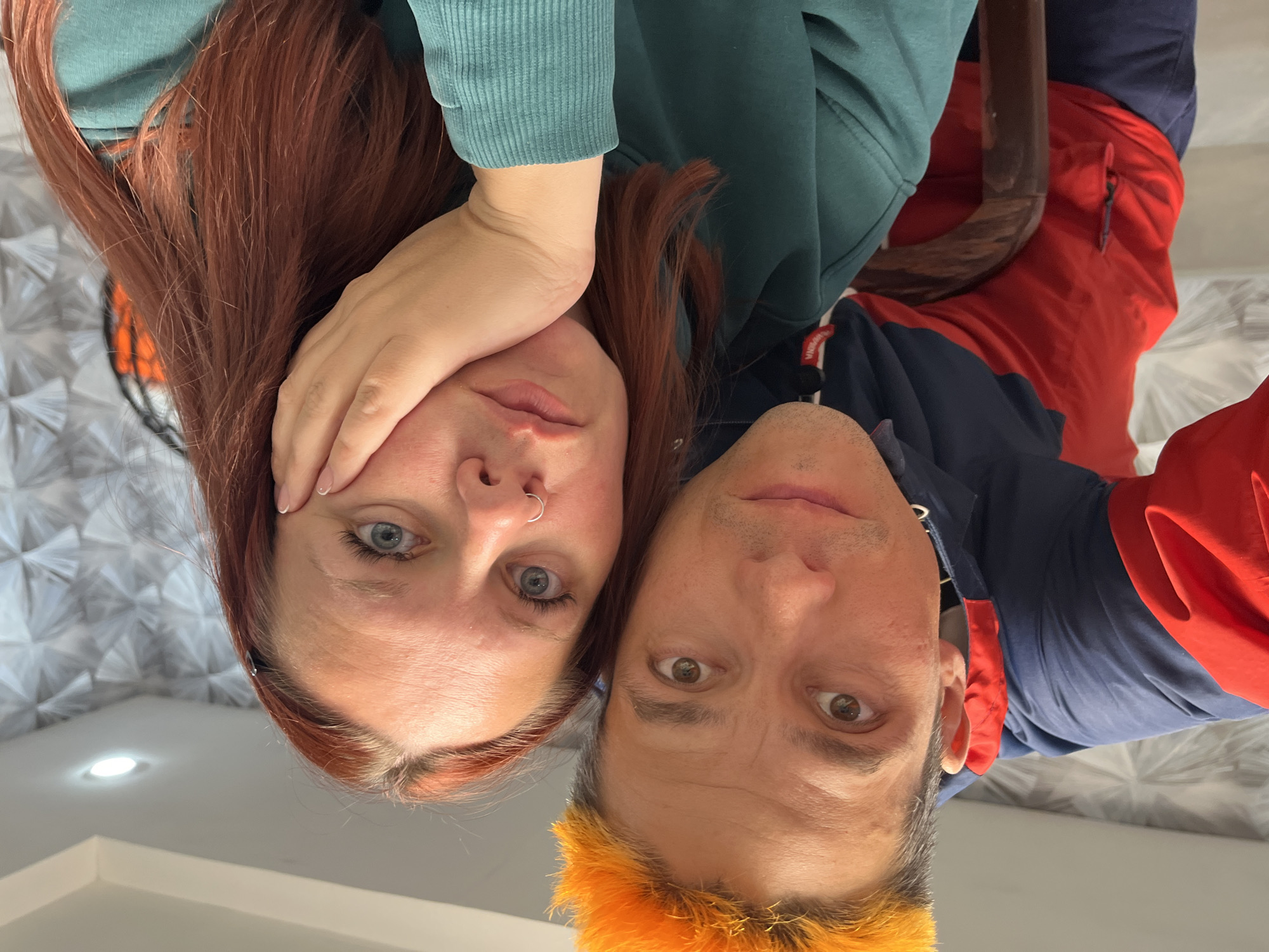 This close-up, low-angle selfie features two individuals, a woman and a man, looking directly at the camera from an upward perspective in an indoor setting in Chaikovsky, Russia.
The woman, on the left, has long reddish-brown hair and blue eyes. She has a silver nose ring on her left nostril and is wearing a teal or dark green ribbed long-sleeved top, likely a hoodie or sweatshirt. Her left hand is raised, partially covering her forehead. Her expression is neutral and appears slightly pensive.
The man, on the right, has short hair with the front and top sections dyed a vibrant orange or yellow. He has brown eyes and light stubble. He is wearing a two-toned top, featuring navy blue and red sections, possibly a hooded jacket or sweatshirt. His expression is also neutral.
The subjects are positioned very close together, with their heads nearly touching. The background is mostly out of focus. Behind the woman, a textured wall or ceiling with a repeating geometric fan-like pattern is visible, along with a bright, recessed ceiling light. Behind the man, a soft furnishing, possibly a bed or sofa with a gray and white patterned cover, can be seen. A dark wooden object, perhaps part of a chair or bed frame, is partially visible between them. On a small white tag on the man's red clothing, faint, difficult-to-discern text is visible, possibly reading "WAVEPNA".
The image captures a casual and intimate moment. The indoor lighting suggests no specific time of day is discernible from the shot itself.
