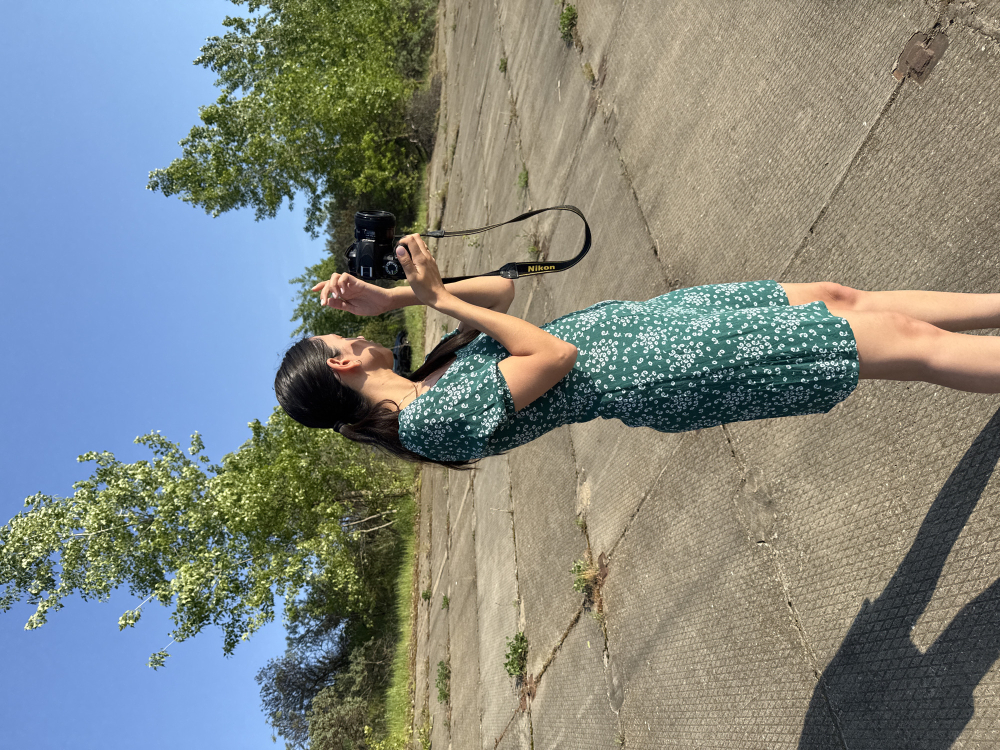 A woman with long, dark hair in a ponytail wearing a short, green and white floral dress is holding a black Nikon camera with a black strap that reads "Nikon" in yellow lettering. She is looking upwards, presumably focusing on a subject to photograph. The background consists of a cracked, concrete surface, likely a paved area, and lush green trees against a clear blue sky. The location is Chaikovsky, Russia.