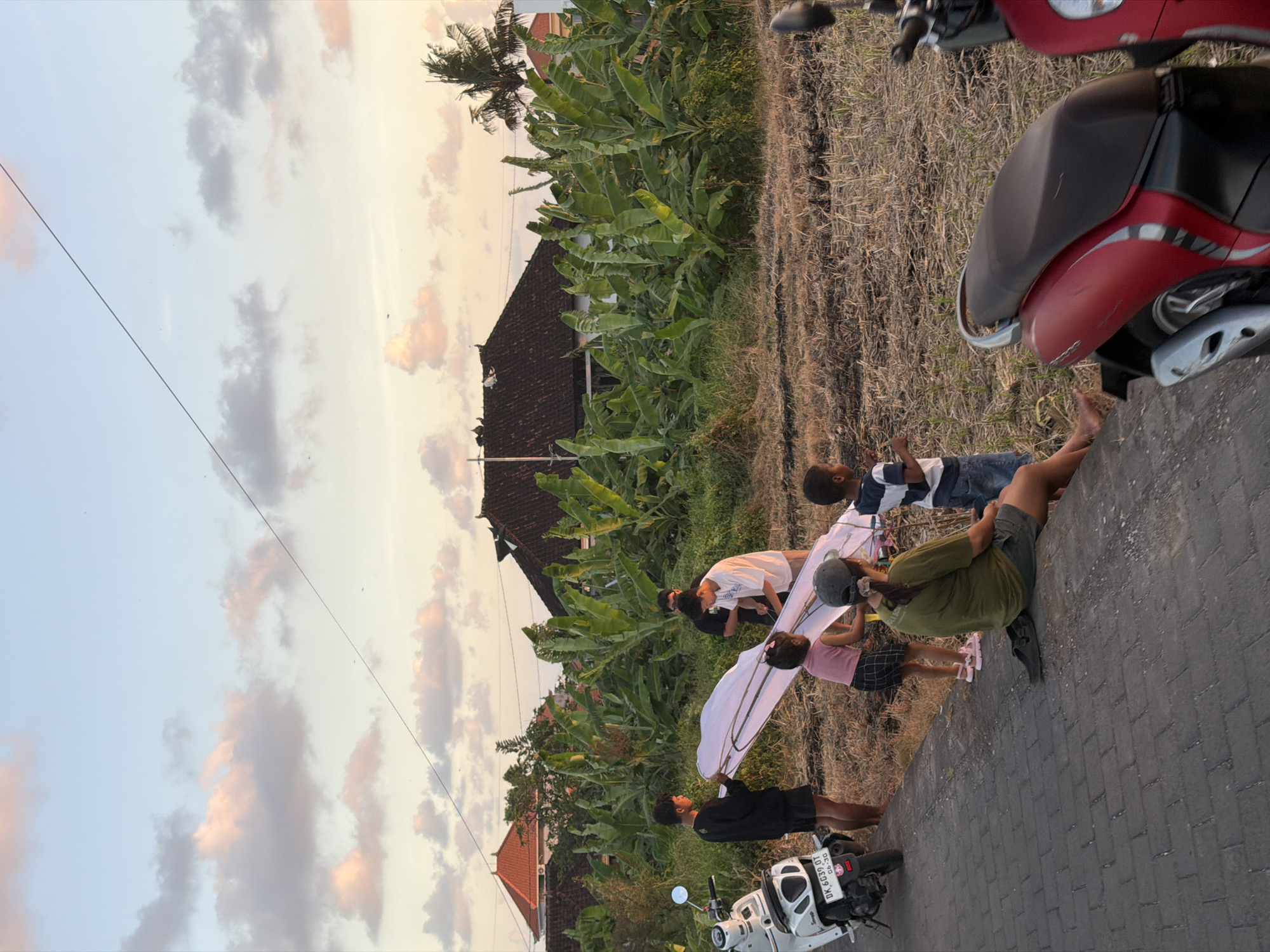 A group of people, including children and adults, are gathered on a paved surface, possibly a road or pathway, in what appears to be a rural setting in North Kuta, Indonesia. The scene is set against a backdrop of lush green banana trees and traditional Indonesian houses with dark tiled roofs. A dry, grassy field stretches alongside the paved area, and a portion of a red and black scooter is visible in the foreground.
The main activity appears to be the assembly or preparation of a large, white kite. Several individuals are interacting with the kite, holding it, and working on its structure. The people are dressed in casual clothing, and some of them seem to be children, indicating a family or community gathering.
The time of day is likely late afternoon or early evening, as suggested by the soft, warm light illuminating the sky, which is filled with scattered clouds tinged with orange and pink hues, characteristic of a sunset. The weather appears to be clear and pleasant. The overall mood of the scene seems relaxed and communal, with people engaged in a shared activity.
The only visible text is on the license plate of the white scooter in the foreground, which reads "DK 6839 GT".