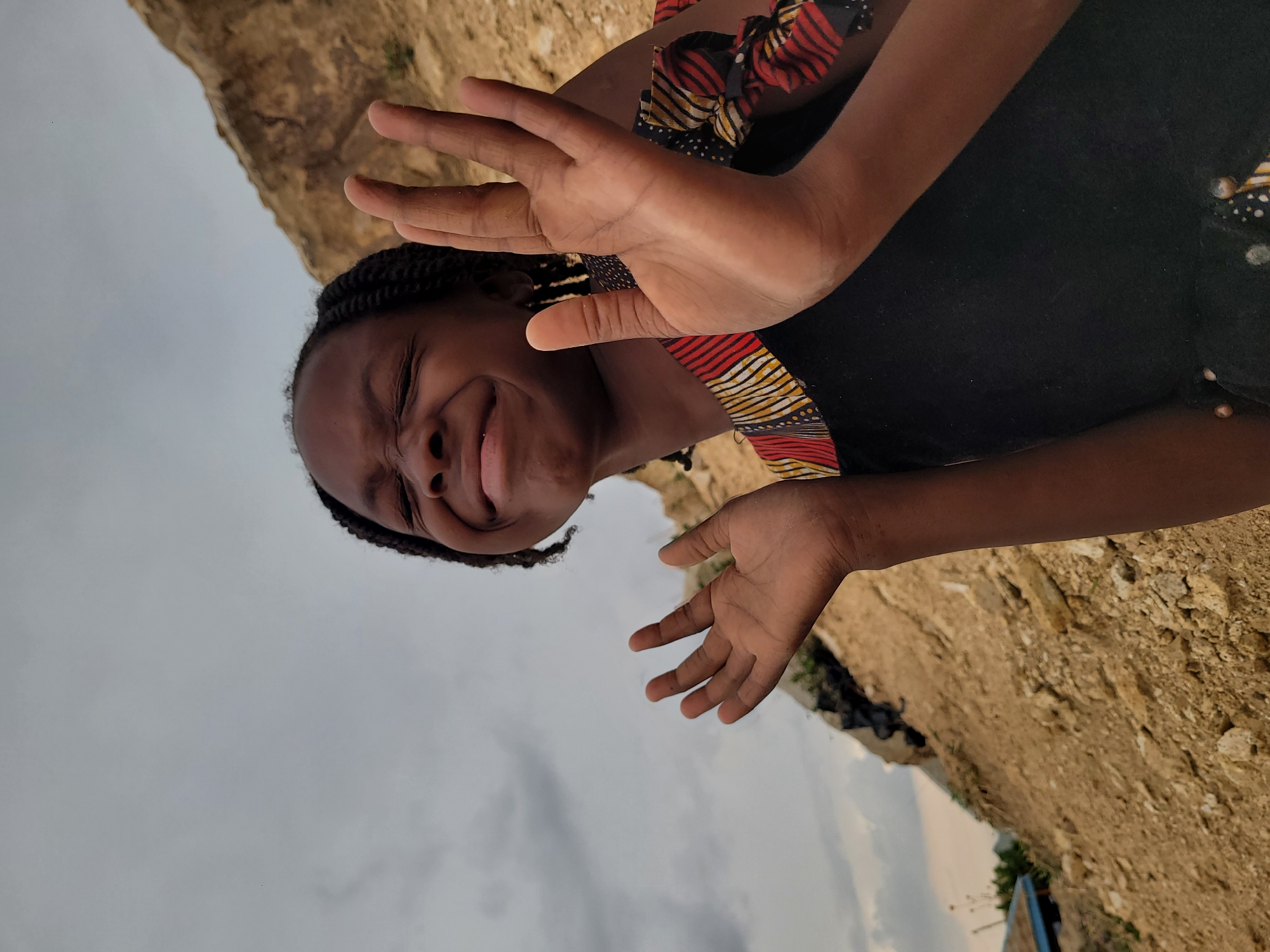 The image captures a young Black girl in Barakin Akawo, Nigeria, posing with her hands raised towards the camera. She is wearing a black dress with a patterned trim in red, yellow, and black. The background shows a sandy, rocky terrain under an overcast sky. The girl appears to be outdoors, with evidence of construction or natural earthworks visible in the background. The scene suggests a location with exposed earth or sand formations, possibly on the outskirts of the city.