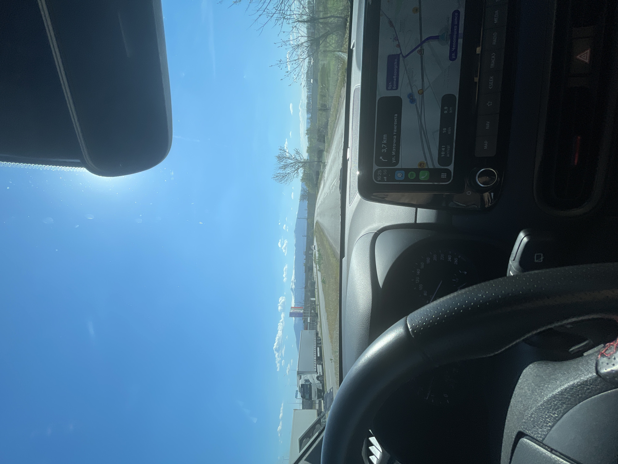 The image depicts a view from inside a vehicle driving on a road, likely a highway, near Sofia, Bulgaria. The sky is clear and blue. The dashboard and steering wheel of the vehicle are visible, along with the rearview mirror. A GPS navigation system on the dashboard displays a route with a destination approximately 3.7 km away on ul. Iztochna Tangenta. The estimated time of arrival is 16:41, with 16 minutes remaining to the destination, and the distance to the next turn is 9.9km. The vehicle is possibly navigating through a commercial or industrial area with visible trucks and commercial buildings along the roadside. There is sparse vegetation and trees on either side of the road.