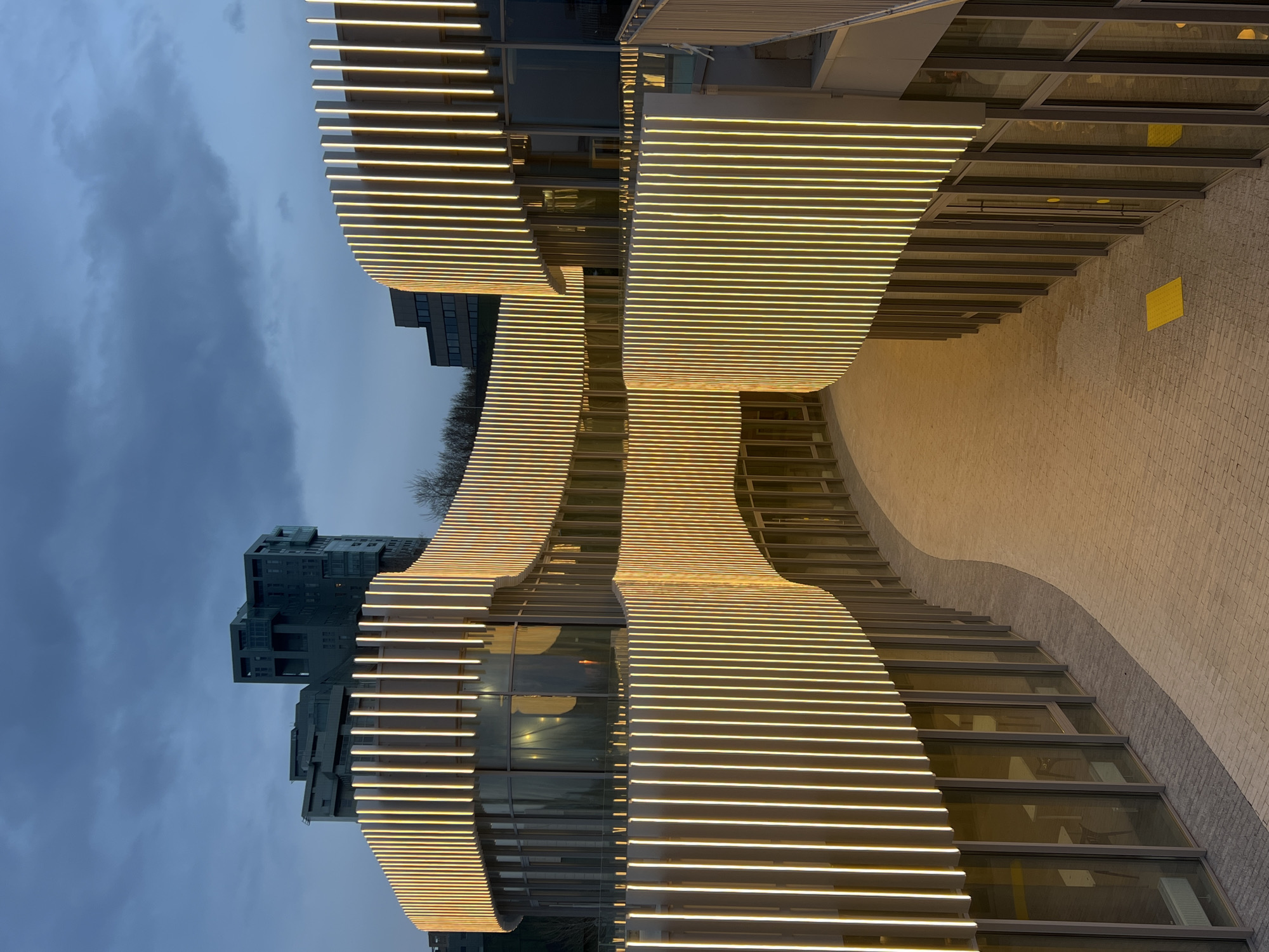 The image depicts a modern architectural structure in Kazan, Russia, identifiable by its distinctive design featuring horizontally aligned, illuminated bars. The building’s facade curves and undulates, creating a dynamic visual effect. Adjacent to the structure, a multi-story building is visible against a cloudy sky, suggesting either dusk or dawn. The ground area appears to be paved, and the overall impression is that of an urban environment with contemporary architectural elements.