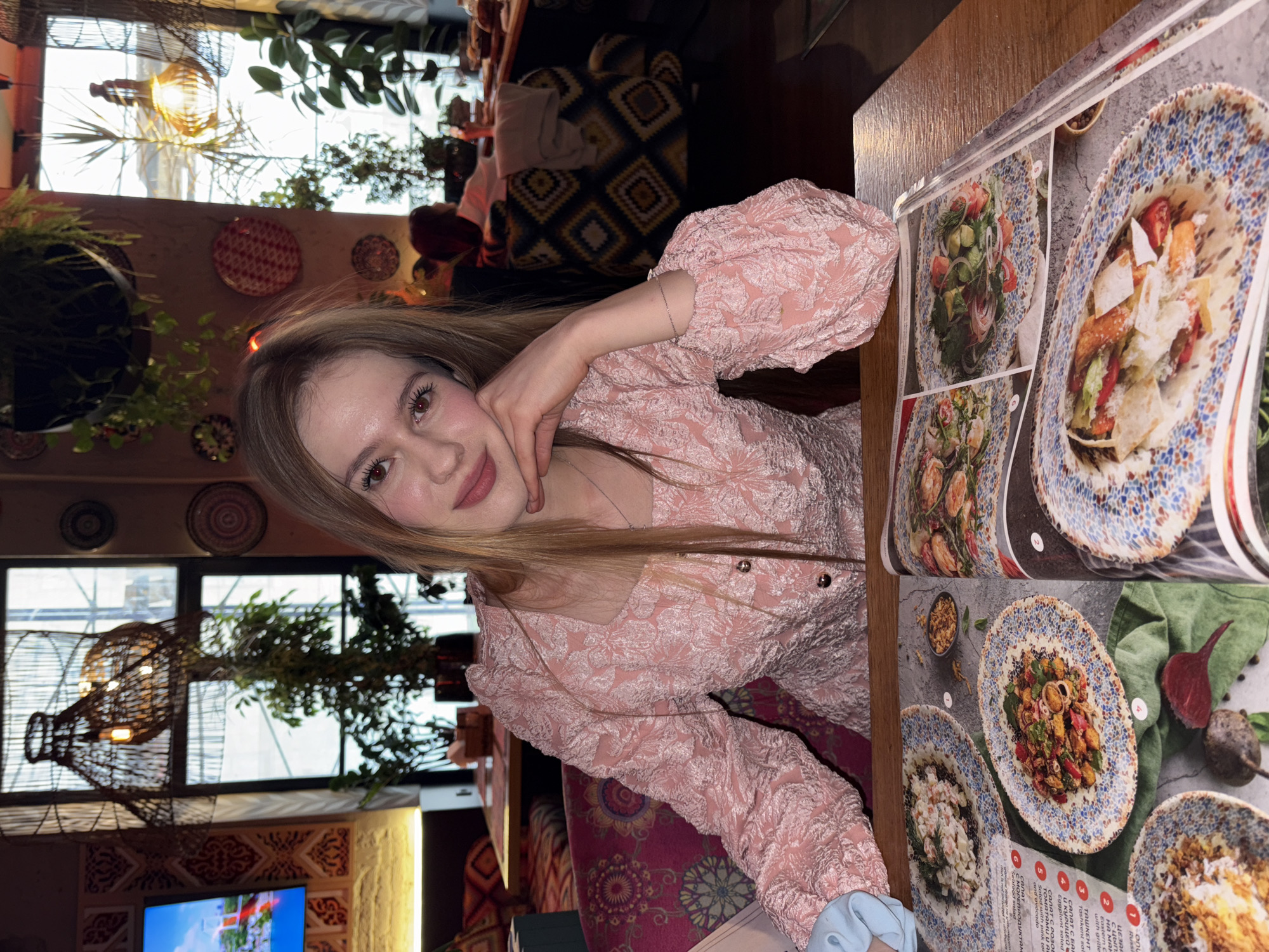 A young woman sits at a wooden table in a restaurant. She is wearing a pink textured top with puffy sleeves and rests her cheek on her hand, smiling at the camera. In front of her is a large menu with photos of various dishes on intricately designed plates. The background is filled with decorative elements including woven baskets, potted plants, and a patterned wall. The image captures a moment of dining in a stylish interior. The OCR text in the image provides details about the menu items, which are salads with various ingredients, including seafood, chicken, and eggplant.