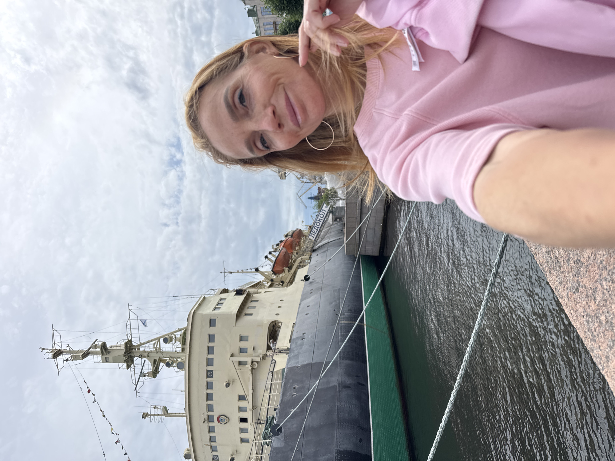 A woman with blonde hair and hoop earrings, wearing a pink sweatshirt, takes a selfie outdoors. Behind her, a large ship, possibly an icebreaker, is docked. The ship is light-colored on top with a dark hull and a green stripe near the waterline. The ship has several windows and appears to be equipped with a crane. In the background, there are buildings and a cloudy sky. The woman is positioned in the foreground, slightly to the right of the frame, with the ship dominating the left side of the image. The scene appears to be set in a harbor or along a waterfront. The ship has the word "ЛЕДОКОЛ" (which translates to "Icebreaker" in Russian) written on its side. This scene is consistent with the location of the Central Naval Museum in Saint Petersburg, Russia, which features the Krassin icebreaker as a monument.
