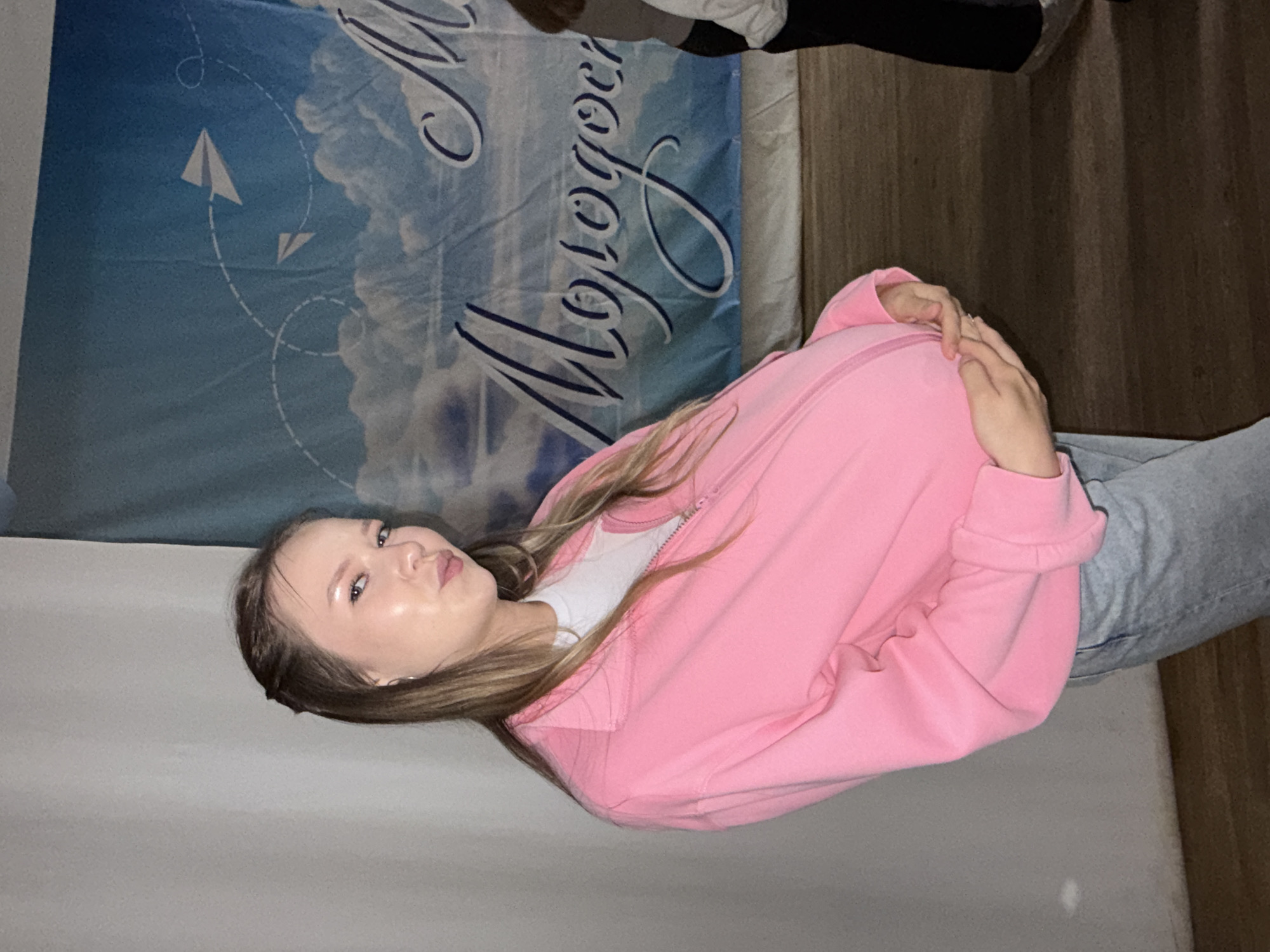 In the city of Chaikovsky, Russia, a young woman stands indoors, posing with her hands cradling her prominent belly, suggesting pregnancy. She is wearing a pink zip-up jacket over a white t-shirt and light-wash jeans. The background features a large banner with a sky motif, clouds, paper airplanes, and Cyrillic text that reads "Молодость" (Molodost), meaning "Youth." The lighting is somewhat dim, creating shadows on the wooden floor.