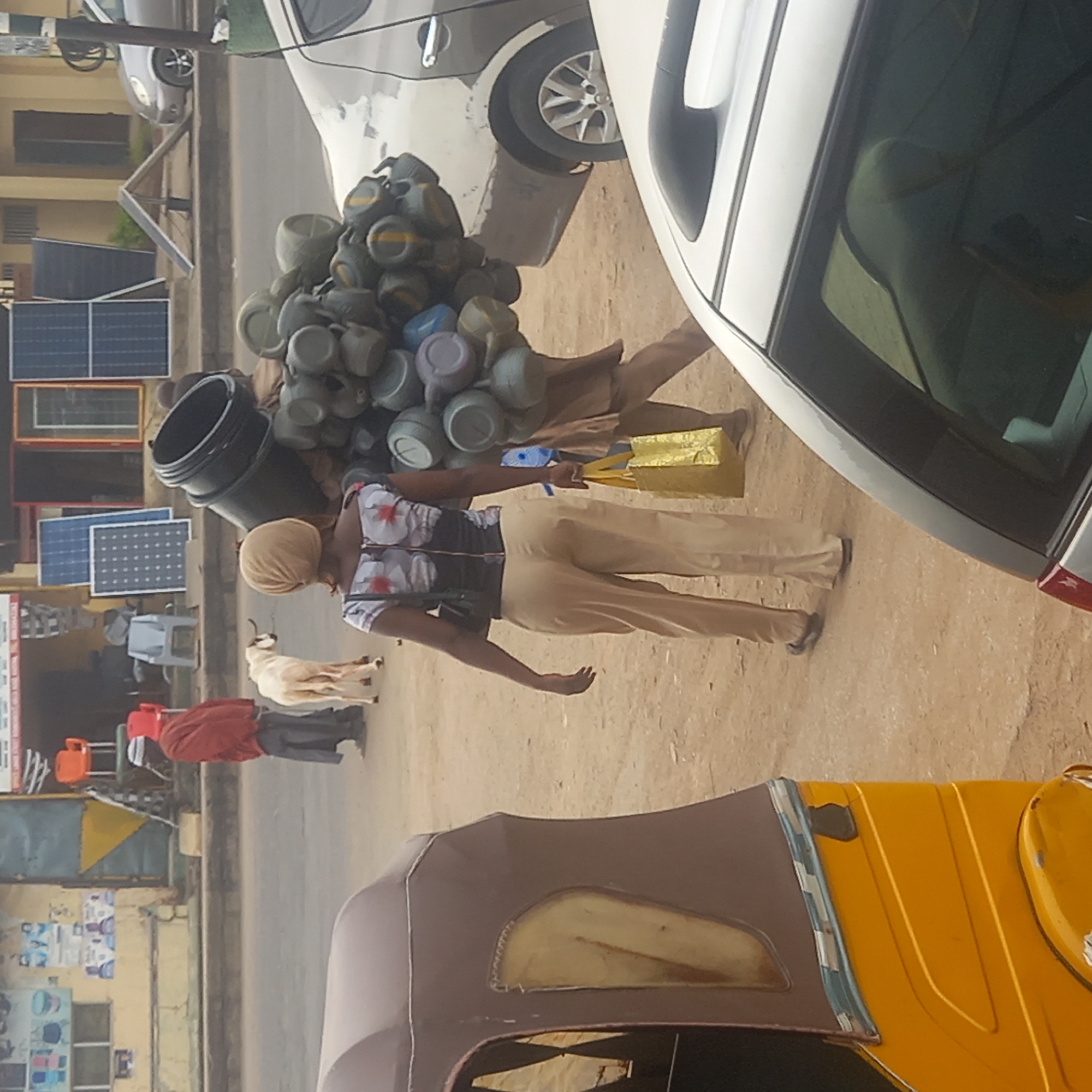 This image captures a bustling street scene in what appears to be Sardauna, Nigeria, during daylight hours under clear, sunny conditions. The perspective is slightly elevated, overlooking the street and its inhabitants.
In the midground, a person is prominently featured, skillfully balancing a large, heaped load of numerous plastic items, resembling mugs or small bowls, on their head. This individual is also carrying a large black bucket on their shoulder and a bright yellow gift bag in their right hand. They are dressed in a short-sleeved top with a floral pattern, light-colored trousers, and a light-colored head wrap. They appear to be walking from the left to the right side of the frame.
Adjacent to the walking individual, a white sedan is visible, either parked or moving slowly. Another grey or silver car is partially seen behind it. To the left of the walking person, a light-colored goat stands on the dusty ground. Further down the street on the left, another person, clad in a red jacket and dark trousers, is standing near some orange chairs.
The background features a row of buildings, which could be commercial establishments or residences. Several solar panels of varying sizes are prominently displayed or stored along the roadside, hinting at a local business dealing in solar energy solutions. The immediate foreground shows the upper portion of a yellow and brown tricycle, commonly known as a Keke NAPEP in Nigeria.
The scene portrays a typical everyday environment, showcasing local commerce, transport, and the integration of livestock within an urban or semi-urban setting. The ground is a mix of dirt and asphalt. No discernible text is clearly legible on any signs or objects due to the image's resolution and distance.