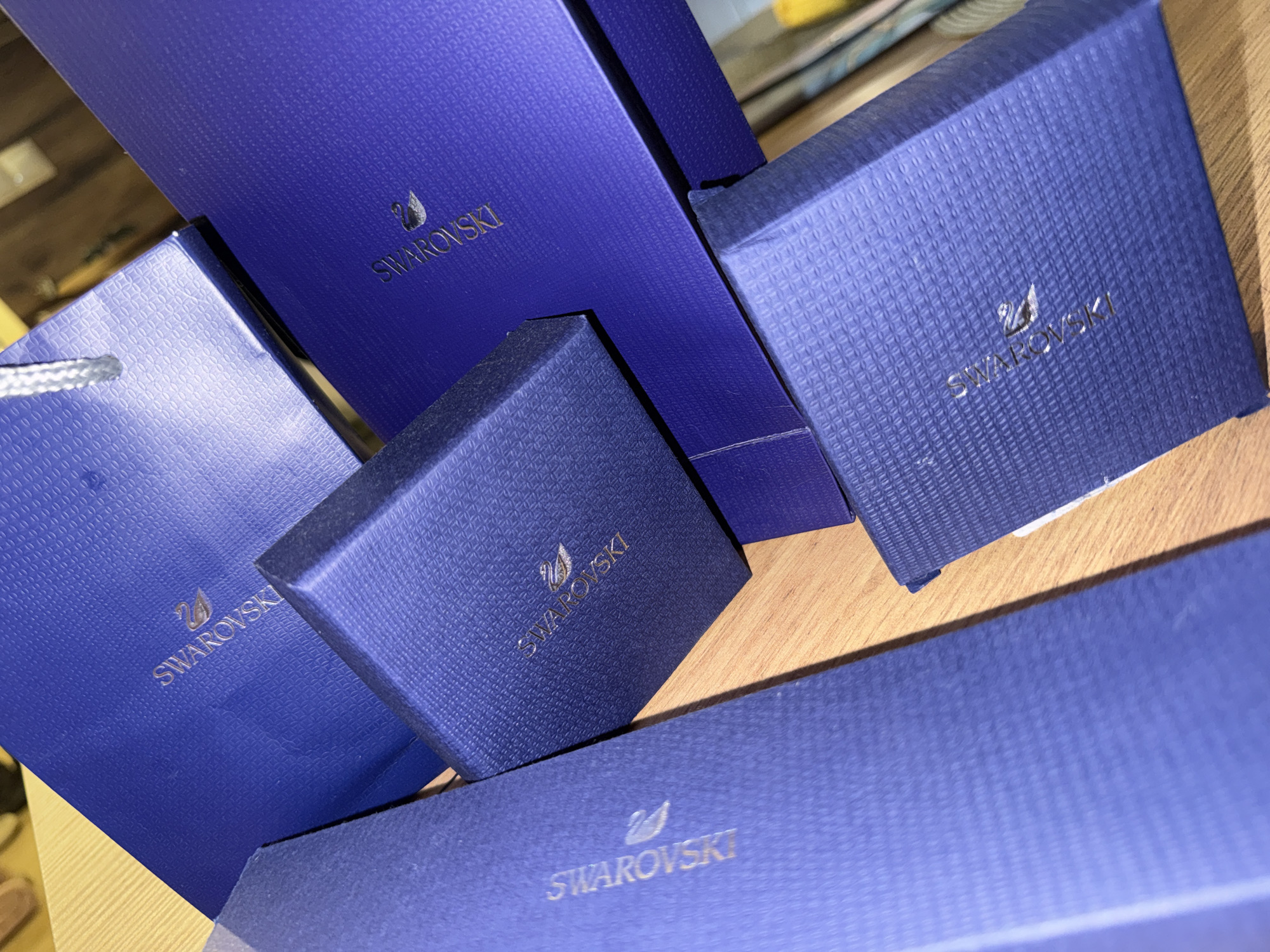The image displays several blue Swarovski boxes and a gift bag arranged on a wooden surface. The boxes have a textured pattern and the Swarovski logo with a swan emblem is embossed in silver on each. The gift bag, also blue, has a silver string handle. The lighting suggests an indoor setting, possibly a retail environment or a home. There are no people or animals visible in the image. The scene is static and focused on the merchandise. The image does not provide any information that can be used to determine its location in the city of Varna, Bulgaria.