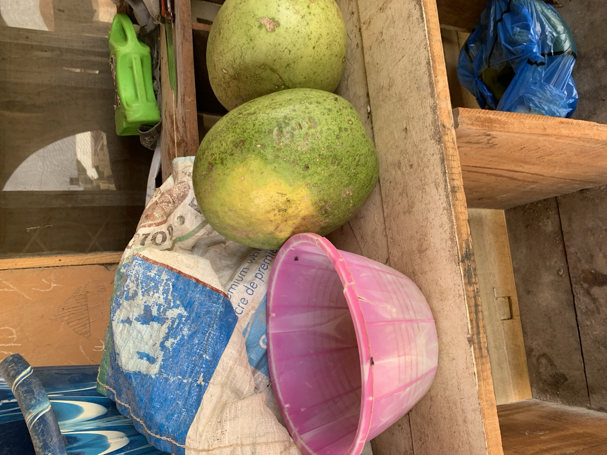 A medium shot shows a wooden stall with a wooden plank shelf at eye level, located outdoors in what is presumed to be the city of Jos, Nigeria. On the shelf, two large, green, melon-like fruits are positioned to the right, one in front of the other. To the left of the fruits is a pink plastic bowl, empty. Behind the bowl and fruits, a large, crumpled bag is visible, half-covered in a blue and white plastic sheet, with some white fabric showing through. The bag appears to be made of woven material and has some printed text in black that is partially obscured by the plastic. In the upper right quadrant of the image, a bright green plastic container with a handle rests on the shelf. A portion of a knife with a red handle is also visible on the shelf to the right of the green container. The stall structure is made of rough, light-colored wood. Below the shelf, a blue plastic bag containing a round object, possibly another fruit, is visible. The ground appears to be made of wooden planks and dirt. The background suggests an open-air environment, with light filtering through a translucent material, creating diffused shadows. The scene is bathed in natural light, indicative of daytime.