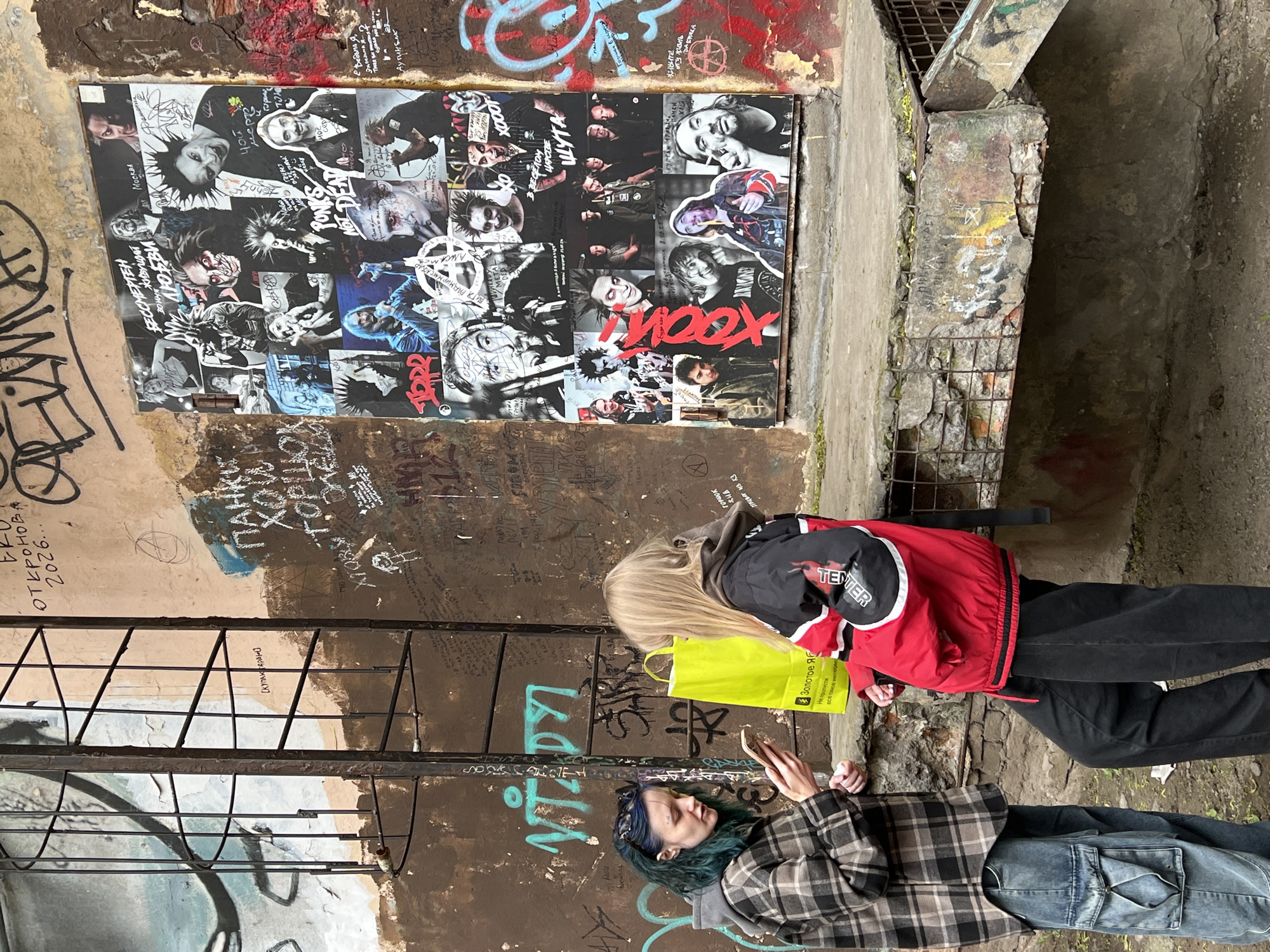 The image depicts an outdoor scene in Saint Petersburg, Russia. Two individuals are present: one woman with long blonde hair is standing, clad in a red and black jacket and black pants, holding a yellow shopping bag. The other woman, with blue hair, is lying on the ground, wearing a plaid shirt and blue jeans, and is looking at a smartphone.
The backdrop is a graffiti-covered wall, with posters and various tags. A significant feature is a poster collage of images of the Russian punk rock band "Korol i Shut" (King and Jester), also known as "КиШ" (Kish). The wall is weathered, showing signs of age and decay. A metal grate is visible on the wall as well. The setting suggests a typical urban environment in Saint Petersburg, possibly a courtyard or alleyway known for its alternative and counter-cultural activities. The image also features the band “Nox”.