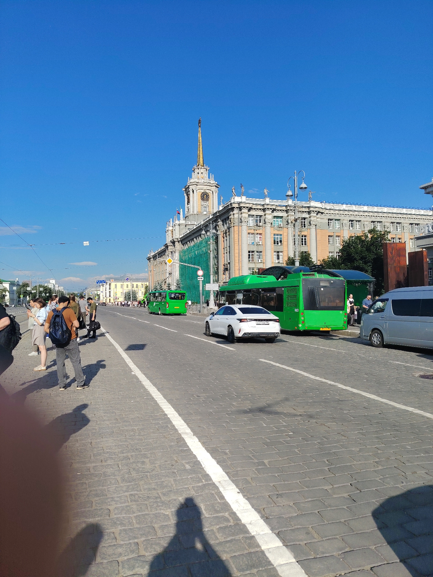 The image depicts a street scene in Yekaterinburg, Russia, on a sunny day. In the background, a prominent building with a spire and clock tower dominates the skyline. The building appears to be a landmark, possibly a government or historical structure, with classical architectural elements and statues adorning its facade. The street itself is paved with cobblestones, and a white line marks the edge of the road.
Several vehicles are present, including a bright green bus, a white sedan, and a silver minivan. People are seen waiting at what appears to be a bus stop. Some are standing on the sidewalk, while others are walking along the street. One individual is wearing a backpack, suggesting they might be a tourist or commuter. Shadows of people are visible on the cobblestone street, indicating the presence of a photographer or a group of people. The sky is a clear, vibrant blue, with no clouds in sight. The overall atmosphere is one of urban activity on a bright summer day.