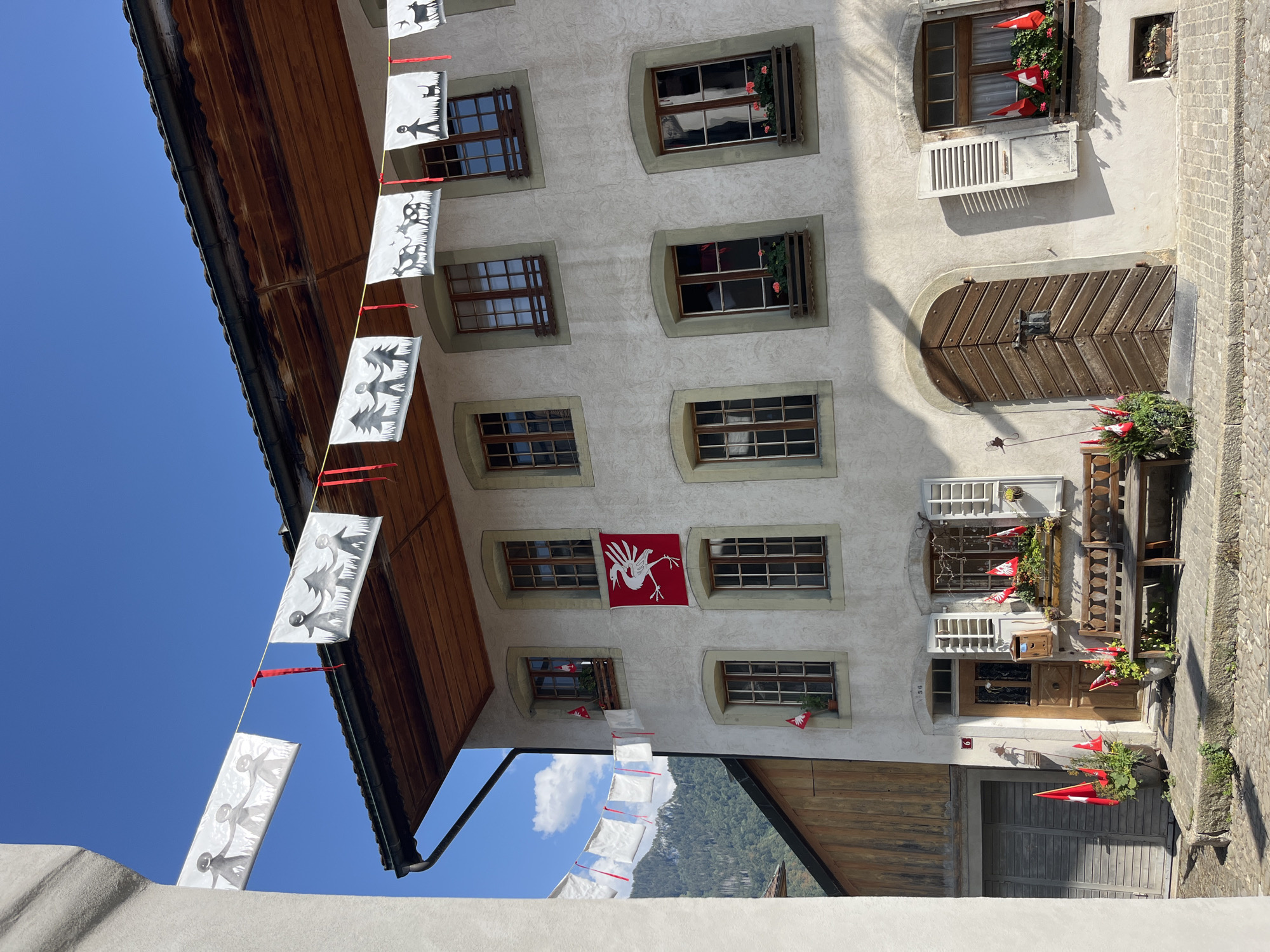 In the city of Epagny, Switzerland, a sunlit, narrow cobblestone street is lined with a building of light-colored stucco. Decorative banners featuring silhouetted figures and animals hang across the street. The building has multiple windows with dark wooden frames and shutters. A red flag with a white bird emblem is displayed prominently on the facade. Swiss flags are placed in window boxes and pots. A wooden bench sits outside a large, arched wooden door, complementing the rustic charm of the setting. The sky is a clear, bright blue.