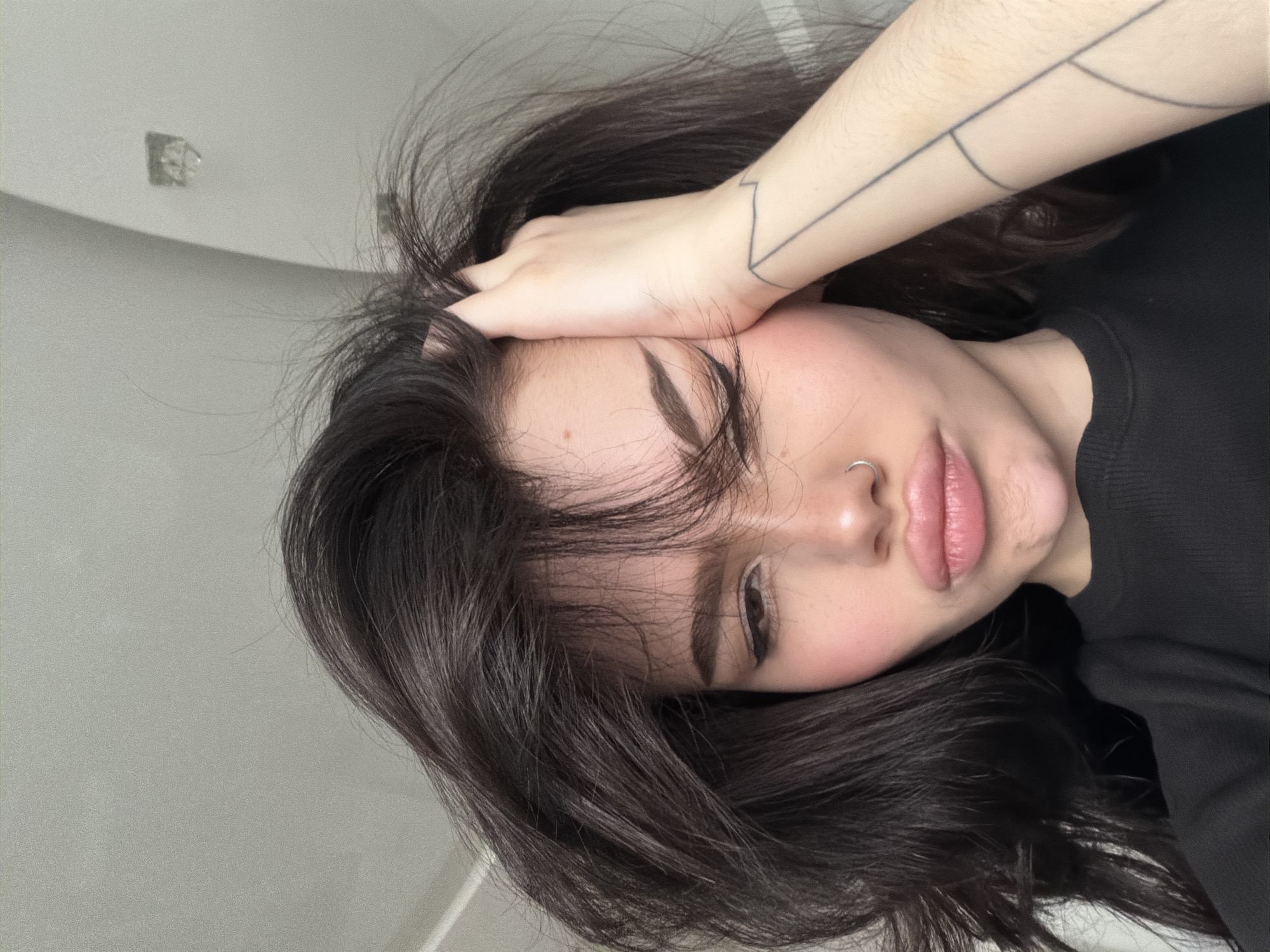 A close-up, rotated image captures a young woman with dark, wavy hair, seemingly lying down indoors. Her head is positioned towards the left side of the frame, with her right arm raised and resting across her forehead, subtly obscuring her eyes and part of her hair. Her fair complexion shows a natural blush on her cheeks, complemented by well-defined eyebrows and full, slightly parted lips. A silver hoop nose ring adorns her left nostril. A prominent black line-art tattoo, featuring geometric or abstract patterns, is visible on her right forearm. She is dressed in a dark, likely black, long-sleeved top. The background consists of a plain, light-colored interior wall or ceiling. In the upper-left portion of the frame, a small, square fixture, possibly an electrical outlet or vent, can be seen on the wall. The lighting is soft and diffused, contributing to a calm and relaxed atmosphere. The woman appears to be resting or in a contemplative state. This indoor scene is situated in Khimki, Russia. There is no visible text in the media file.