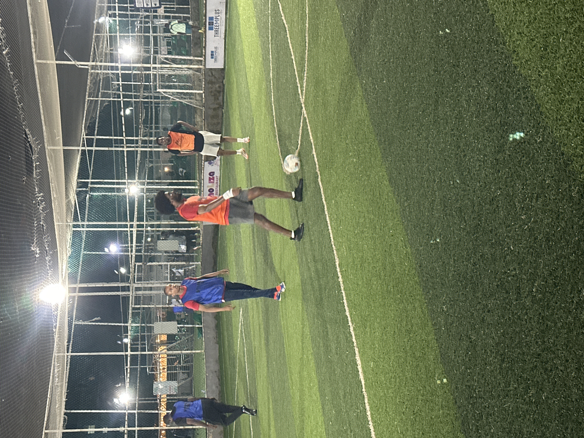 An image shows a group of men playing soccer on an artificial turf field at night. The field is illuminated by overhead lights. The men are wearing athletic clothing, some with bibs. A soccer ball is visible on the ground in the foreground. In the background, there are fences and buildings. The scene is likely an outdoor sports facility.