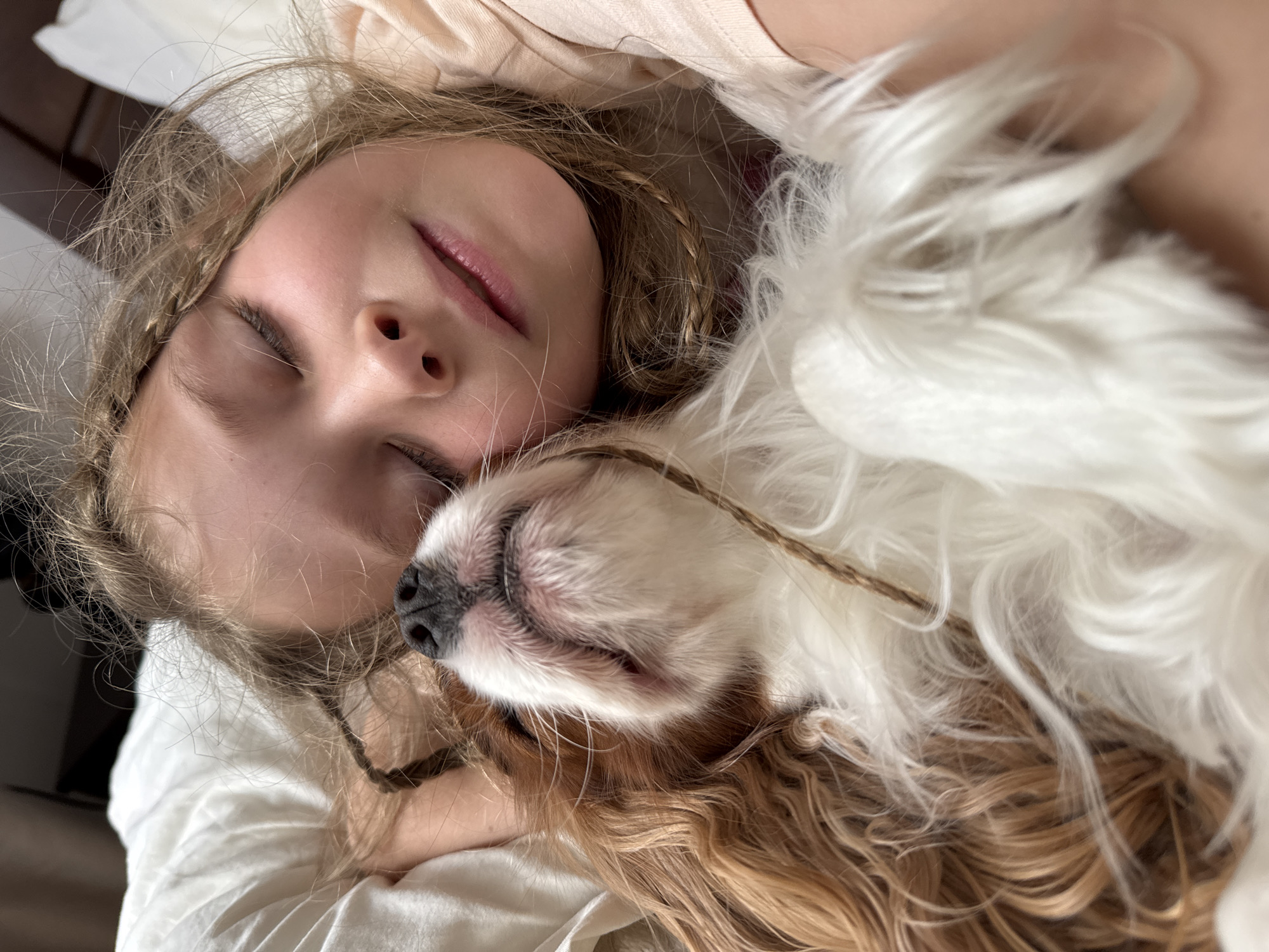 A close-up, angled photograph captures a serene moment between a young girl and a small dog, both appearing to be asleep or resting. The girl, positioned on the left side of the frame, has light-colored hair with visible braids, and her eyes are gently closed. Her face shows a peaceful expression, with slightly parted lips. She is wearing light-colored clothing, possibly a soft top or pajamas.
Cuddled closely beside her face on the right is a small, long-haired dog, predominantly white with patches of light brown or tan fur. The dog's eyes are also closed, and its snout, featuring a dark nose, is nestled near the girl's cheek. Both subjects are surrounded by soft, white bedding or fabric, suggesting an indoor setting like a bed or couch. The lighting is soft and natural, implying daytime. The overall mood of the image is one of deep comfort, affection, and tranquility. There is no visible text.