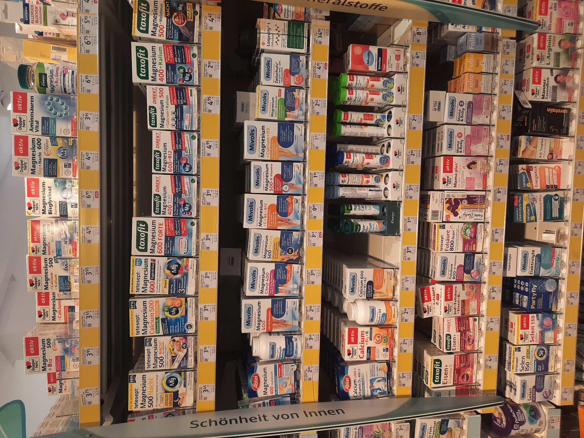 The image shows a shelf display in a drugstore or pharmacy in Rosenheim, Germany. The shelves are filled with various health and wellness products, primarily dietary supplements, with a strong emphasis on magnesium-based products. Brands like Doppel Herz, Mivolis, Taxofit, and Abtei are prominently featured. There are also products for vitamins, calcium, iron, and other supplements. The overall impression is one of a well-stocked retail environment focused on personal health and well-being. The products are neatly arranged in their packaging, with price tags visible on many of them. The lighting suggests an indoor retail setting. No people are visible in the image.