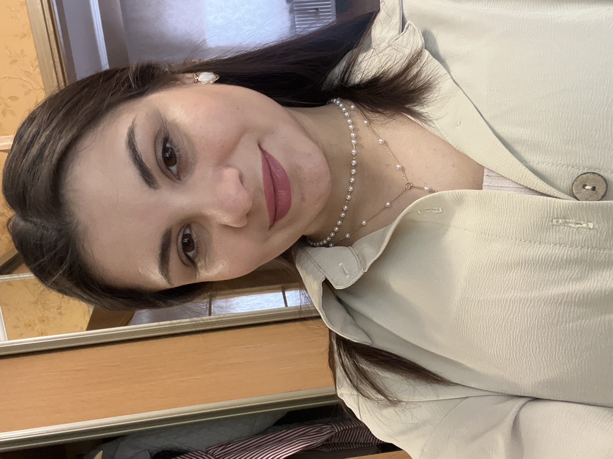 A medium-close-up, waist-up, selfie-style portrait of a woman with dark brown hair, brown eyes, and fair skin. She is wearing a light beige collared shirt with a pearl necklace and a gold pendant. She has on pink lipstick and small white earrings. The background is blurred but shows a wooden closet door to the left, and a wall with patterned wallpaper to the right, with a window visible. The lighting is dim, and the image is slightly grainy.