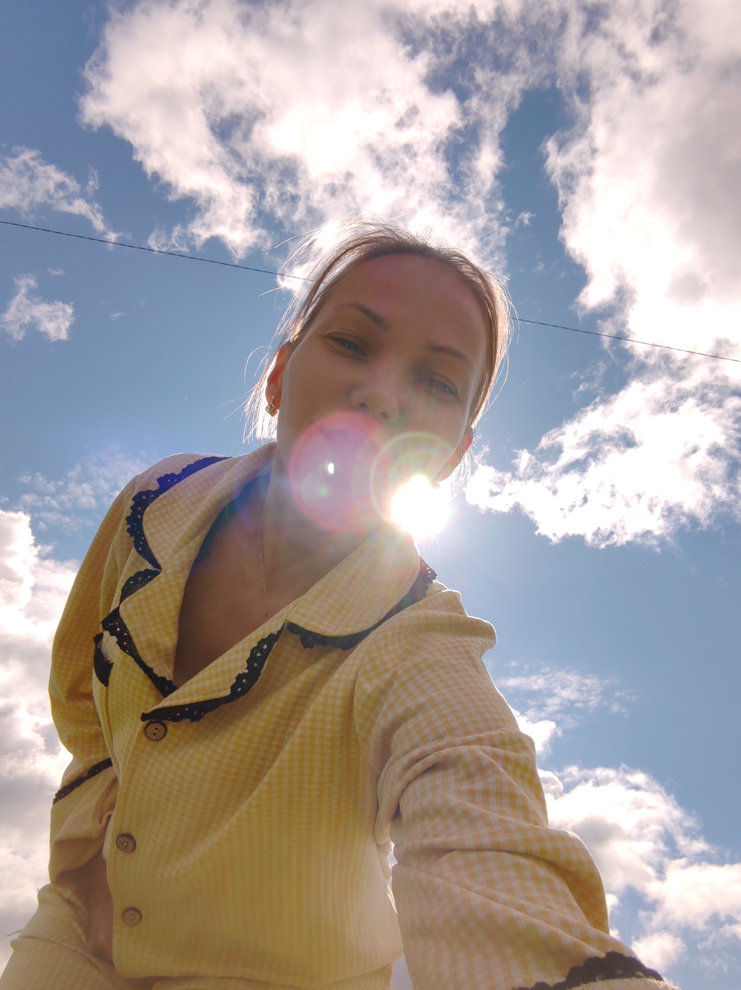 A low-angle shot captures a woman wearing a yellow and white checkered pajama set with black lace trim around the collar and cuffs. The woman is looking up at the camera, with the sun directly in front of her, creating a lens flare that obscures her face. Her hair is light brown and pulled back, and she is wearing small gold earrings. The background is a bright blue sky filled with fluffy white clouds, suggesting it is daytime and the weather is fair. The image appears to be a selfie taken outdoors. The setting is identified as Чухлэм, Russia. No visible text is present in the image. The overall mood is bright and perhaps playful, given the selfie perspective and the cheerful colors.