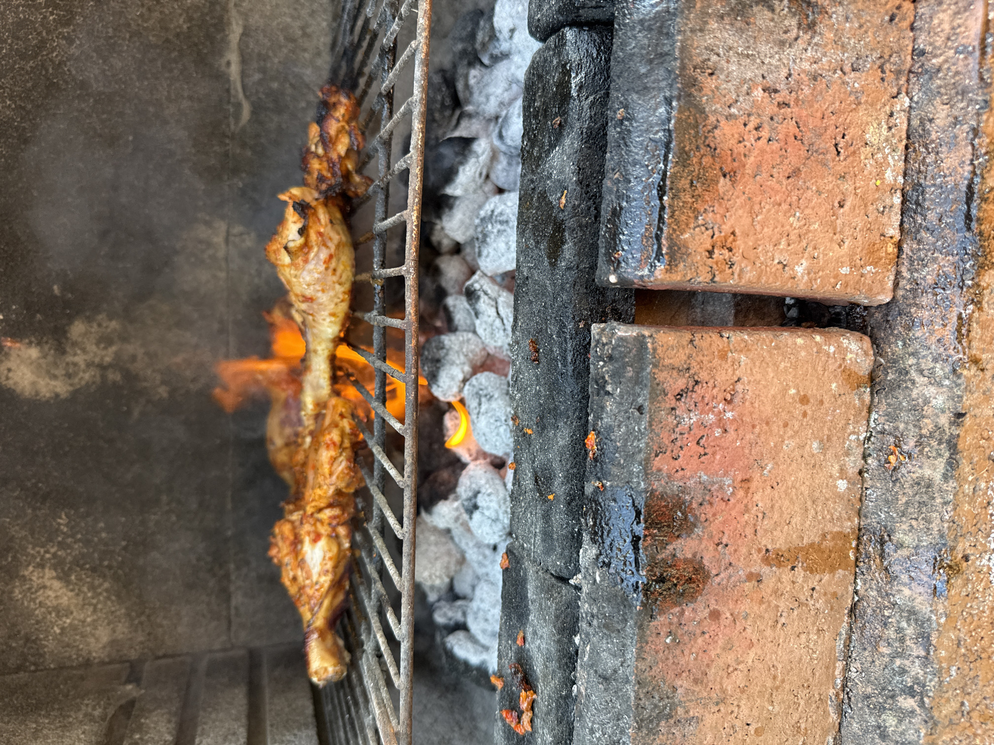 The image shows chicken legs on a grill over hot coals. The grill is inside a brick barbecue pit. The chicken appears to be seasoned with spices and is being cooked over an open flame. The background is a textured stone wall. The scene is likely a backyard or outdoor cooking area. There are no people visible in the image. The location in Rendsburg, Germany is not indicated in the image or context.