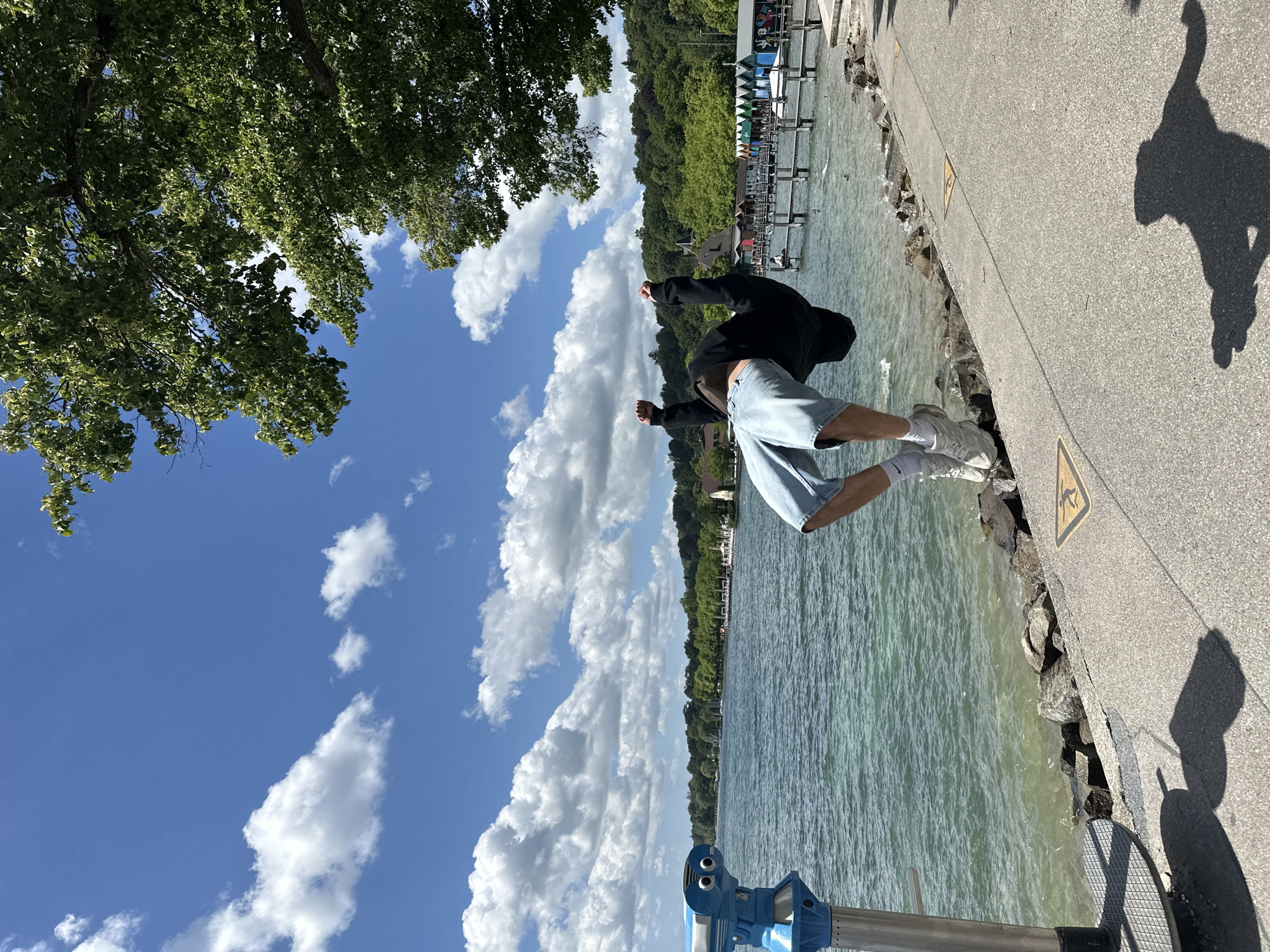 A young person is captured mid-air performing a backflip next to the Starnberger See in Germany. The individual, dressed in a black hoodie and denim shorts, is positioned over the rocky shoreline with the lake's calm, greenish-blue water extending into the background. A scenic view of the lake is visible, with trees lining the opposite bank and a pier with colorful boat houses in the distance. The sky is a vibrant blue, dotted with fluffy white clouds, and a large tree branch overhangs the scene from the upper right corner. A blue tourist telescope is positioned on the left side of the frame, and the shadow of the person performing the flip is cast onto the concrete pathway.