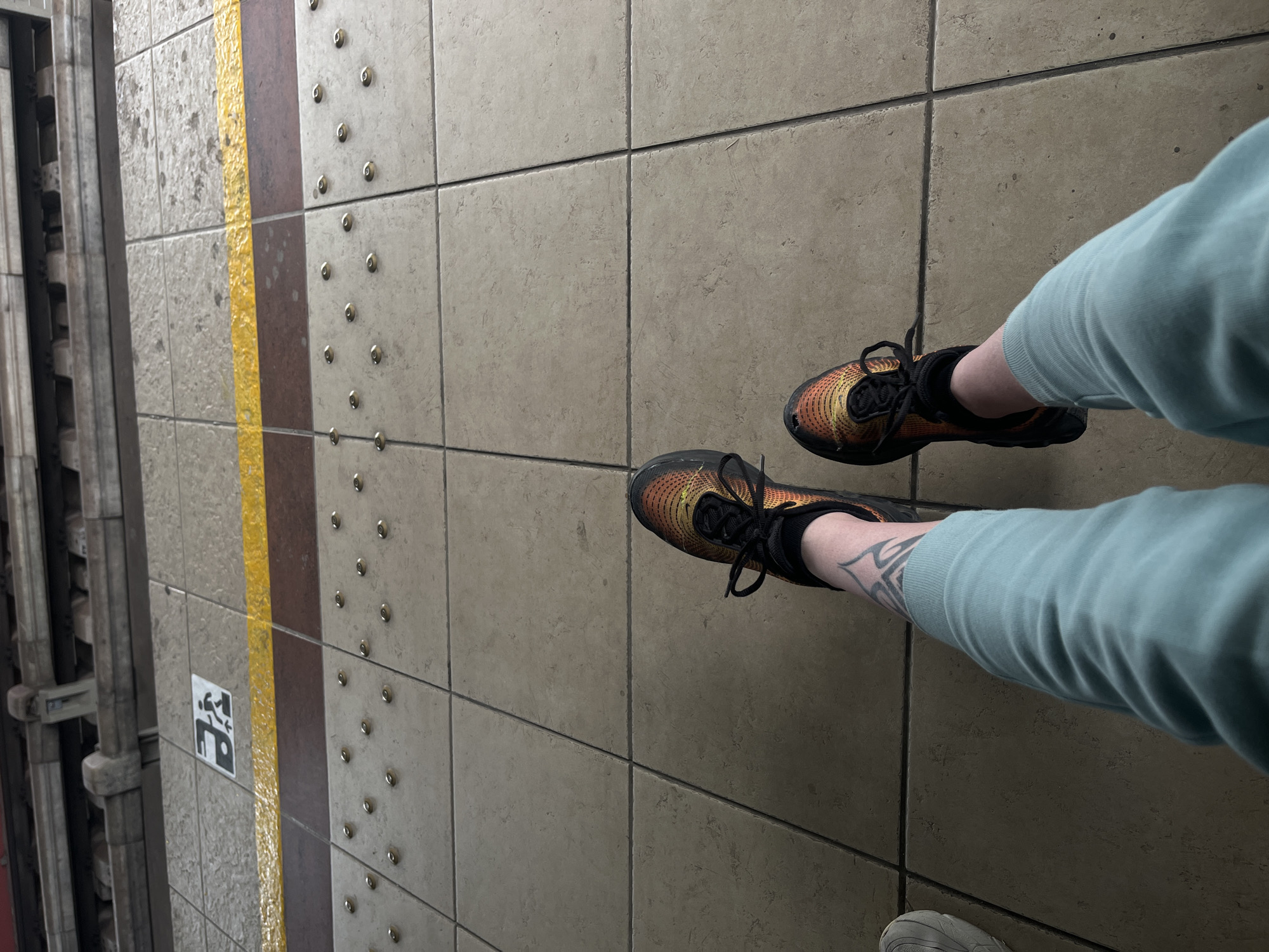 The image shows a person standing on a subway platform. The platform is tiled with a beige tile, marked with a yellow line and brown tiles. A subway door is on the left side of the image. The person is wearing orange and black sneakers and light blue sweatpants. There is a tattoo on their ankle. The location is not in Sofia, Bulgaria.