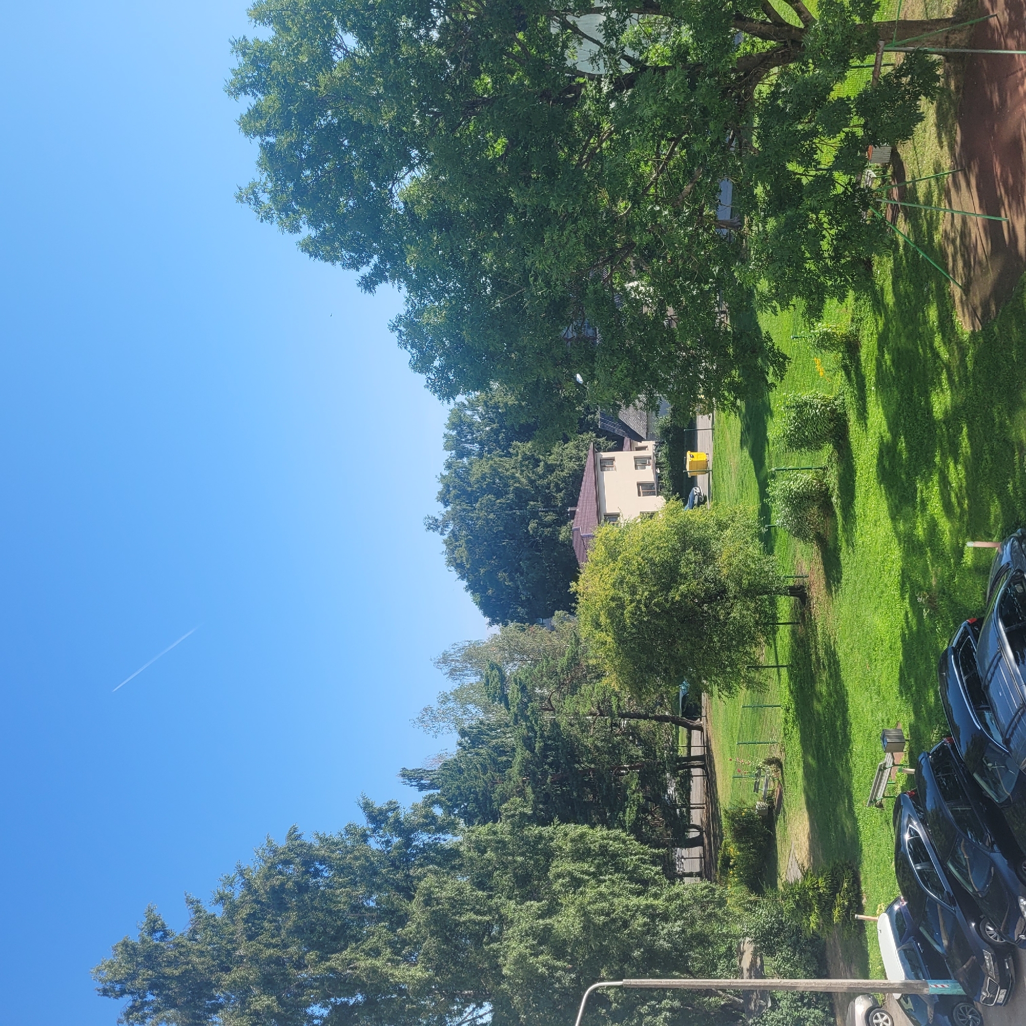 The image shows a residential area in Jūrmala, Latvia, on a sunny day. The sky is clear blue with a faint contrail from an airplane. In the foreground, several parked cars are visible, including black sedans and SUVs. A lamppost stands to the left of the cars. Beyond the cars, a well-maintained green lawn stretches out, dotted with trees and bushes. A few park benches and a trash can are placed on the grass. In the middle ground, a two-story building with a brown roof is visible, surrounded by trees. The background is filled with lush green trees and foliage, indicative of a natural or park-like setting. The scene suggests a peaceful, everyday environment in a suburban or semi-urban area.
