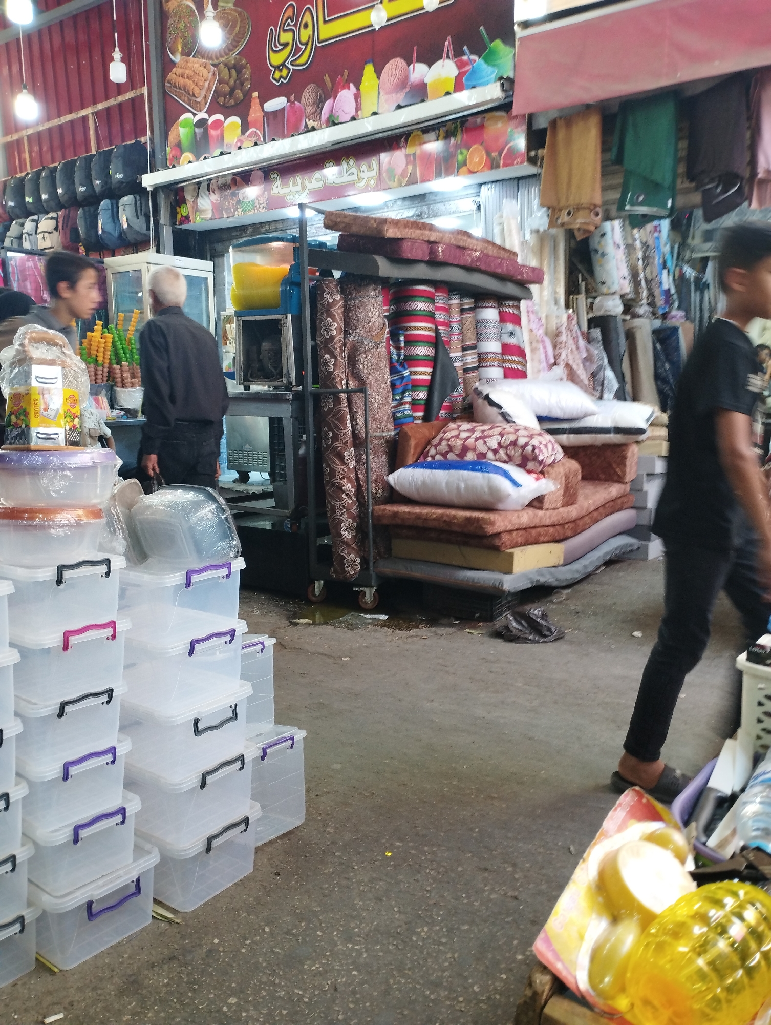 The image depicts an indoor market stall, possibly located in Baqa'a camp, Jordan. A young male, wearing a black t-shirt and black pants, walks towards the right side of the frame. In the foreground, stacks of clear plastic storage containers with colored lids are arranged. Behind them, a man with gray hair, wearing a black long-sleeved shirt and dark pants, stands near a display of colorful ice cream cones. Further back, a refrigeration unit is visible, with a large container of yellow liquid inside. The stall also features a display of textiles, including patterned rolls of fabric and hanging garments. Overhead, promotional signs with images of ice cream and drinks are mounted. The overall environment appears to be a bustling marketplace.