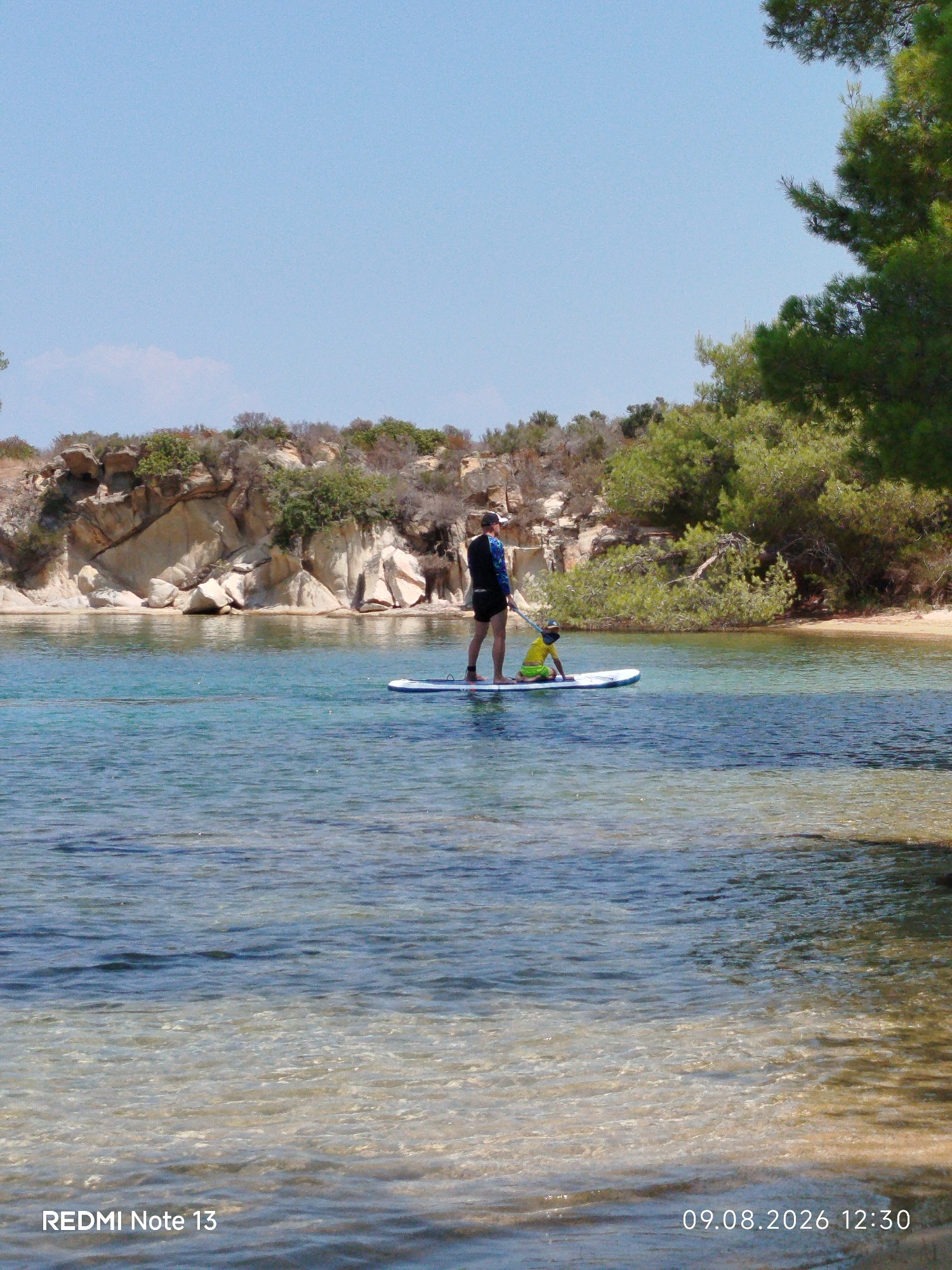 Two people are paddleboarding on a calm, clear body of water. One person, an adult, is standing on the paddleboard, wearing a dark shirt with blue sleeves and dark shorts, and a cap. The second person, a child, is kneeling on the paddleboard, wearing a yellow shirt and green shorts, with a hat. The paddleboard is white with blue accents. The water is shallow near the shore, revealing the sandy bottom, and deepens to a darker blue further out.
In the background, a rocky, tree-covered hillside slopes down to the water. The rocks are light brown and grey, with patches of green vegetation growing on them. Further back, dense green trees create a natural backdrop. The sky is clear and blue. The location is a coastal area within the Sithonia Municipal Unit in Greece, likely a sheltered bay or cove. The image has a timestamp in the lower right corner indicating "09.08.2026 12:30". In the lower left corner, the text "REDMI Note 13" is visible.