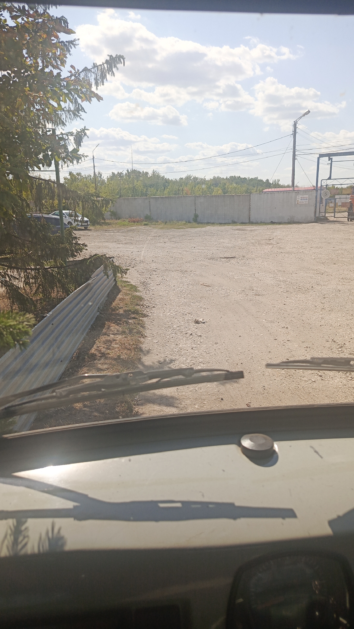 The image depicts an outdoor scene on a sunny day, as viewed from inside a vehicle. In the foreground, the dashboard and windshield wipers of the vehicle are visible. The exterior environment includes a dirt or gravel lot, a grey concrete wall, a corrugated metal fence, and trees. Beyond the wall, more trees and a blue sky with white clouds are visible. A street light on a pole is also present, as are power lines. To the right, a metal gate is partially open, revealing a structure beyond. On the wall, near the gate, there is a sign with text and a symbol. Two cars and a bicycle are parked near the left side of the concrete wall. The scene appears to be an industrial or semi-industrial area, possibly a parking lot or a service area, located in or near the city of Золотая Степь, Russia.
