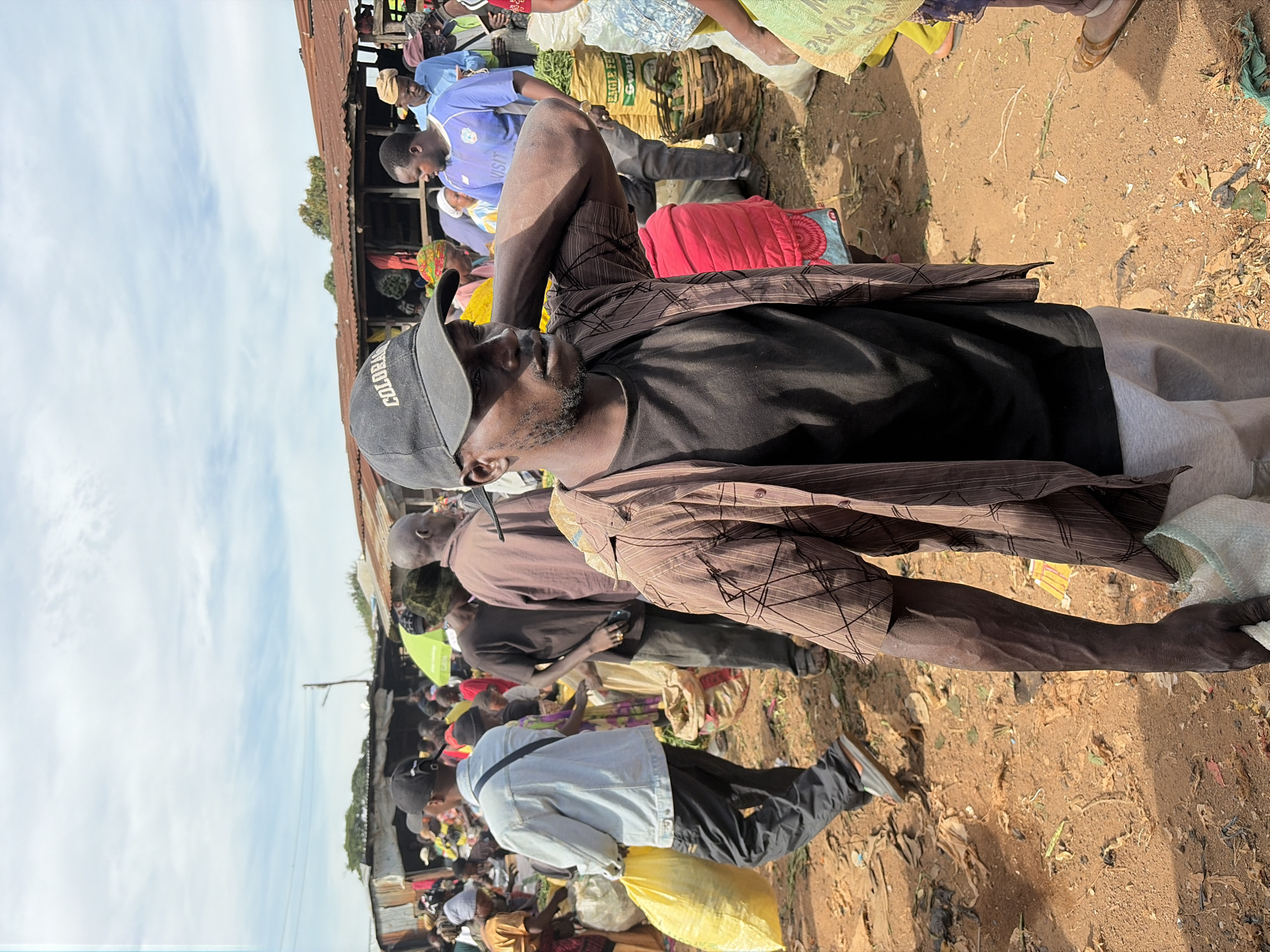 A busy outdoor market scene in Gura Top, Nigeria, unfolds under a cloudy sky. The foreground is dominated by a man wearing a black t-shirt, a brown plaid-patterned button-up shirt, light grey sweatpants, and a black baseball cap with "COLORADO" embroidered on it. He is holding a white sack in his right hand and has his left arm behind his head, appearing to scratch his neck.
Behind him, other market-goers navigate the crowded space. To his left, a man in a light blue denim jacket and dark pants carries a large yellow sack. Further back, individuals in casual attire, including a man in a blue t-shirt with a logo, are visible amongst stalls and baskets piled with produce. The ground is covered in reddish-brown dirt, scattered with debris like dried leaves and plastic. The market stalls are rustic, with corrugated metal roofs and wooden structures, indicative of a local, informal marketplace. The overall atmosphere suggests a vibrant, active community engaged in commerce.
