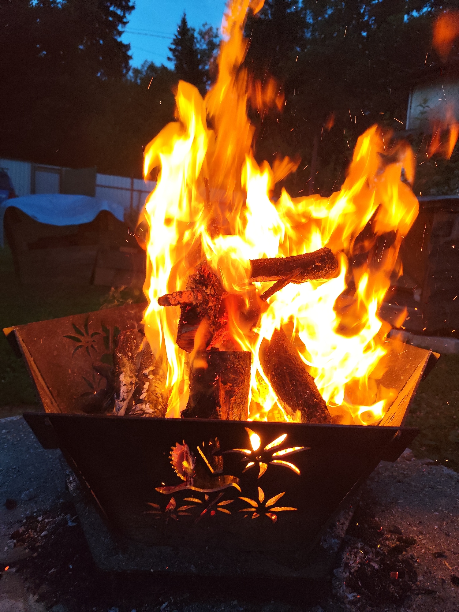 The image shows a close-up view of a fire pit in Kolpakovo, Russia. The fire pit is made of dark metal and has decorative cutouts of palm trees and a bird. Inside the fire pit, logs are burning, producing bright orange flames and sparks. The background is dark, with silhouetted trees and a twilight sky. A blue tarp-covered object and a fence are visible in the distance. The ground around the fire pit is covered in ash and embers.