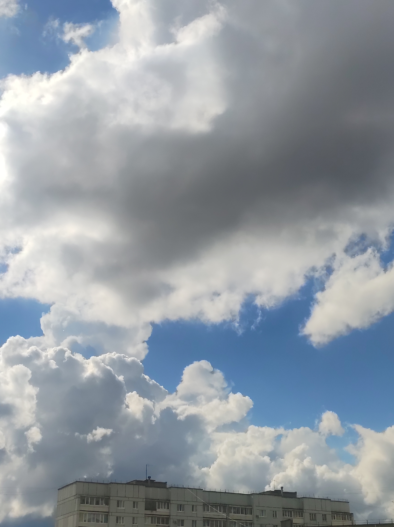 The image captures a dramatic sky dominated by a dynamic interplay of clouds and blue patches, with the top of a building visible at the bottom.
The upper portion of the frame features a vast expanse of heavy, dark grey cumulus clouds, suggesting a significant weather system or thick cloud cover. Below these, and extending across the middle and lower sections, are voluminous, bright white cumulus clouds, brilliantly illuminated by sunlight, creating a stark contrast against the darker clouds and occasional clear blue sky. Streaks of deep blue sky are visible in various areas, particularly towards the top left and middle right, peeking through the cloud formations. The strong sunlight highlights the edges and textures of the white clouds, giving them a three-dimensional, almost mountainous appearance.
At the very bottom of the frame, the top floors and roofline of a multi-story, light-colored building are visible. The building appears to be a residential or administrative block, characterized by numerous uniform windows and a flat roof adorned with what appear to be antennas or vents. The architecture suggests a style common in towns like Chaikovsky, Russia, fitting the provided context.
The scene depicts daytime weather conditions, likely midday or afternoon, given the intensity of the sunlight on the clouds. The combination of dense, dark clouds and bright, sunlit patches indicates a day of shifting weather, possibly with scattered showers or the clearing after rain. No specific activity or people are visible, and there is no discernible text within the image.