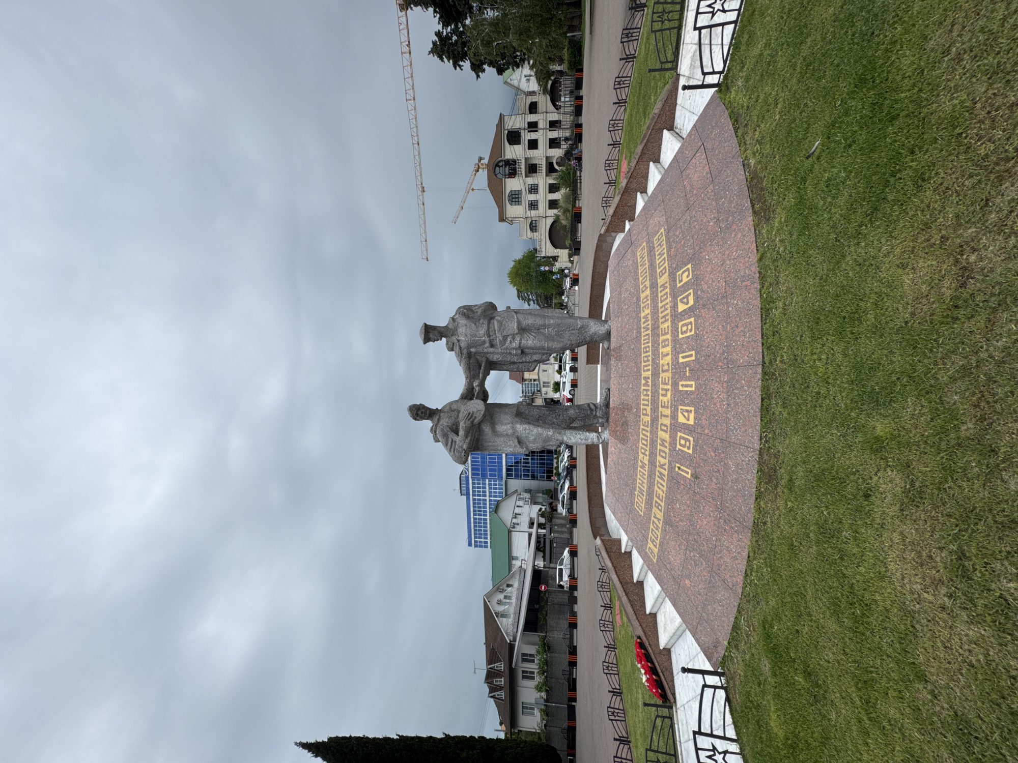 This image shows the "Anchor and Cannon" monument located in the city of Sochi, Russia. Two statues of soldiers, likely commemorating World War II, stand on a circular, red-and-brown platform. The platform contains inscription in Russian. The area surrounding the platform is covered in green grass, and a metal fence encloses it. Buildings of various architectural styles are visible in the background, including a multistory building and some smaller residential houses. There are also two construction cranes visible in the distance, indicating ongoing construction activity. The sky is overcast, suggesting a cloudy day.