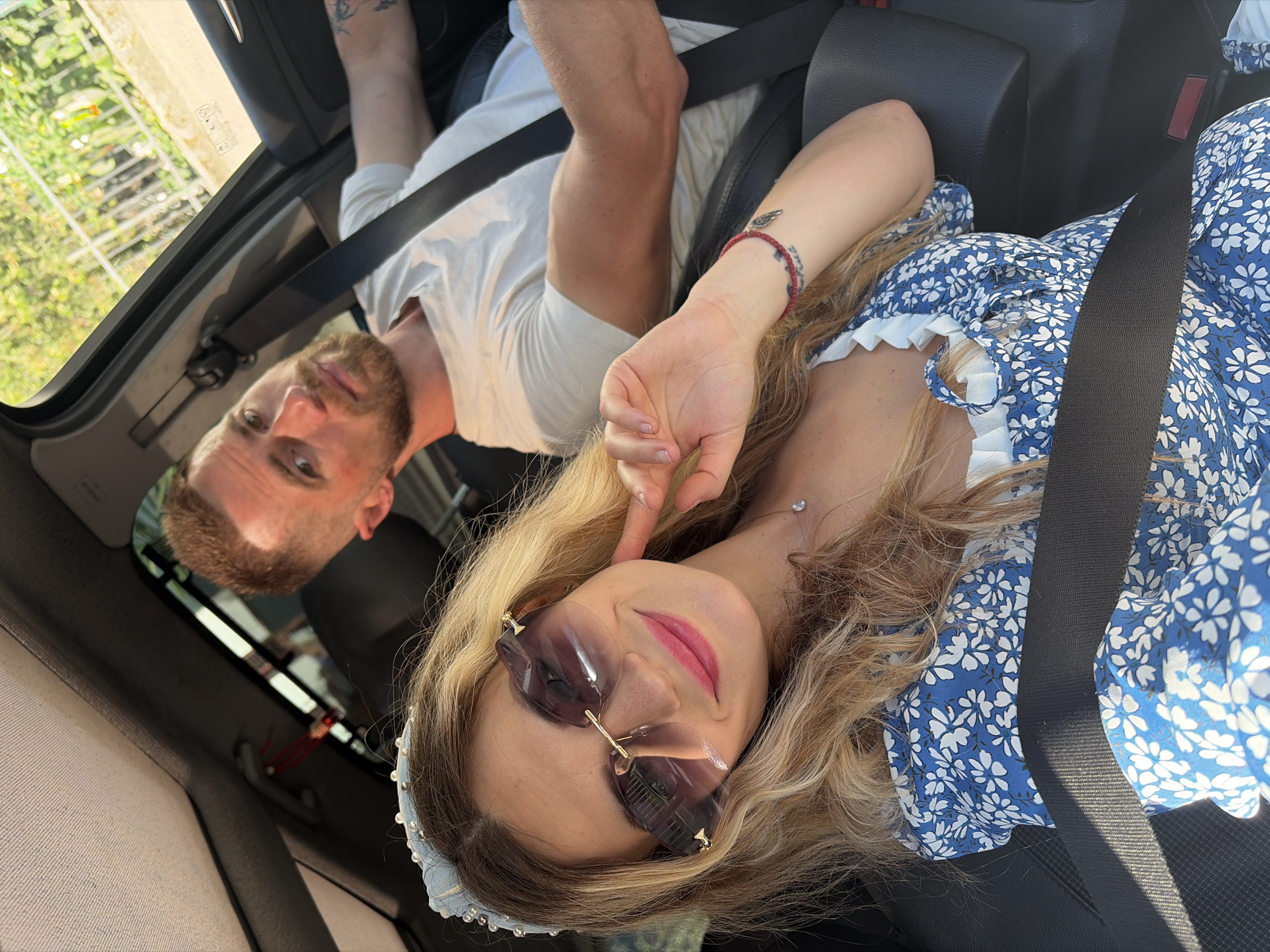 A blonde woman with a pearl headband, large designer sunglasses, and a blue and white floral dress is in the passenger seat of a car. She is smiling and touching her cheek. A man with short brown hair and a beard, wearing a white t-shirt, is driving the car. The car interior is visible, including the dashboard, steering wheel, and seatbelts. The exterior of the car shows trees and greenery through the windows, suggesting a location with vegetation. The image appears to be taken during the daytime, with sunlight illuminating the scene. The man has a tattoo on his arm.