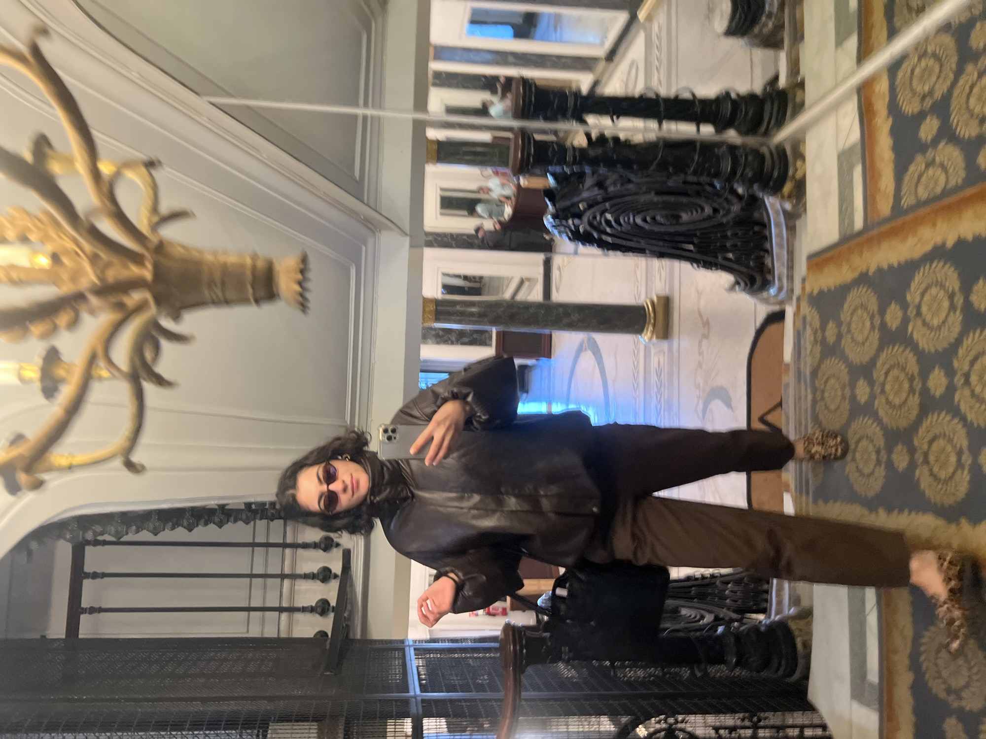 A person stands in front of a mirror. The person is wearing sunglasses, a brown jacket, and brown pants. The person is holding a phone and looking at the camera. Behind the person is a metal railing. Above the person is an ornate chandelier. The mirror reflects a hallway with pillars and a rug with a floral pattern. Based on the ornate architectural details and decor, it is possible that this photo was taken in Paris, France.