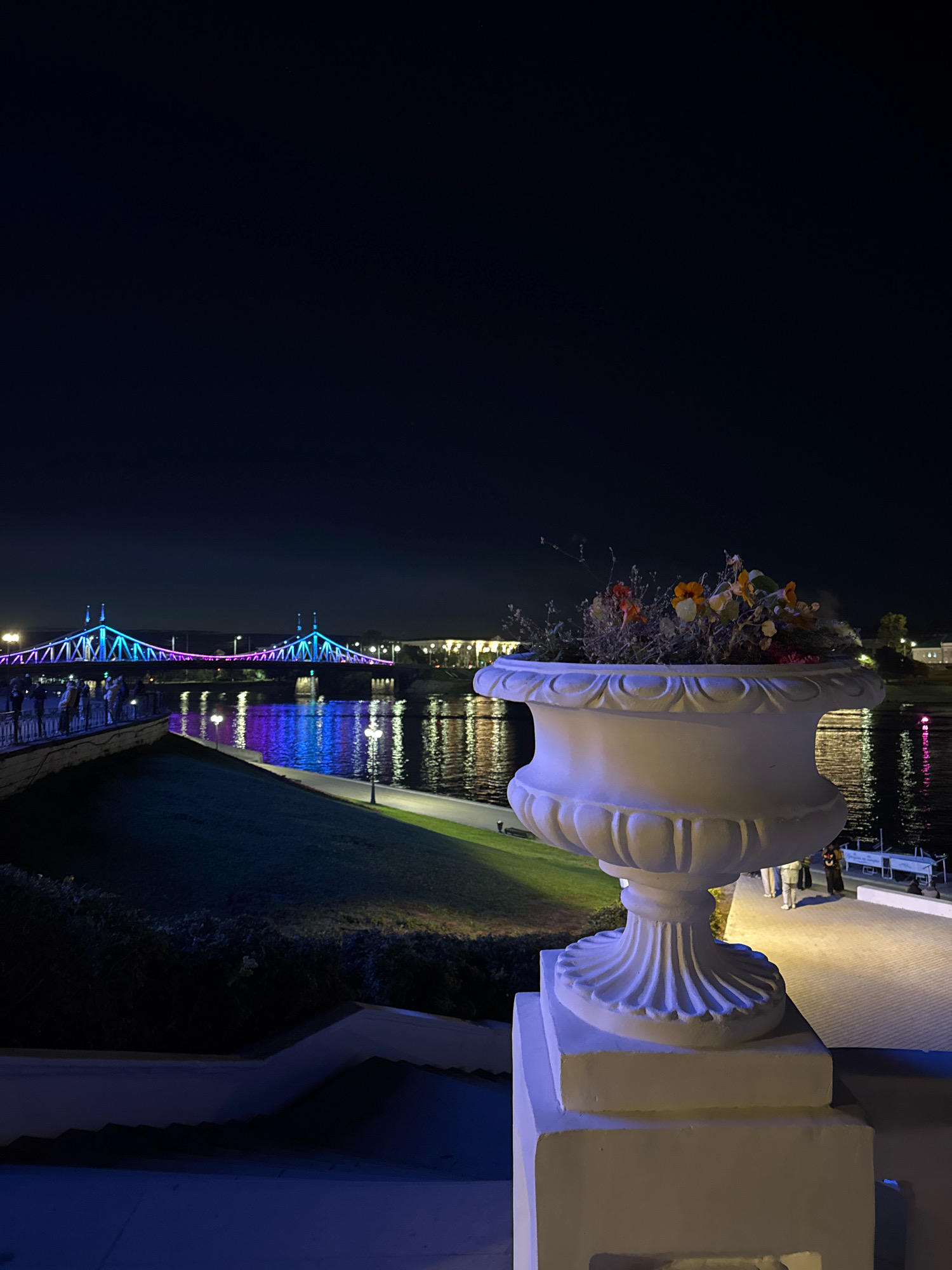 The image captures a nighttime scene in Tver, Russia, along the embankment of the Volga River. In the foreground, a large, ornate white planter with dry flowers sits on a pedestal. Beyond the planter, a paved walkway is illuminated, with several people walking along it. To the left, a grassy slope leads down to the river. The river itself reflects the colorful lights of a bridge in the distance, which is illuminated with blue and pink hues. People can be seen gathered on the embankment near the bridge. The overall atmosphere is one of a lively, illuminated urban waterfront at night.