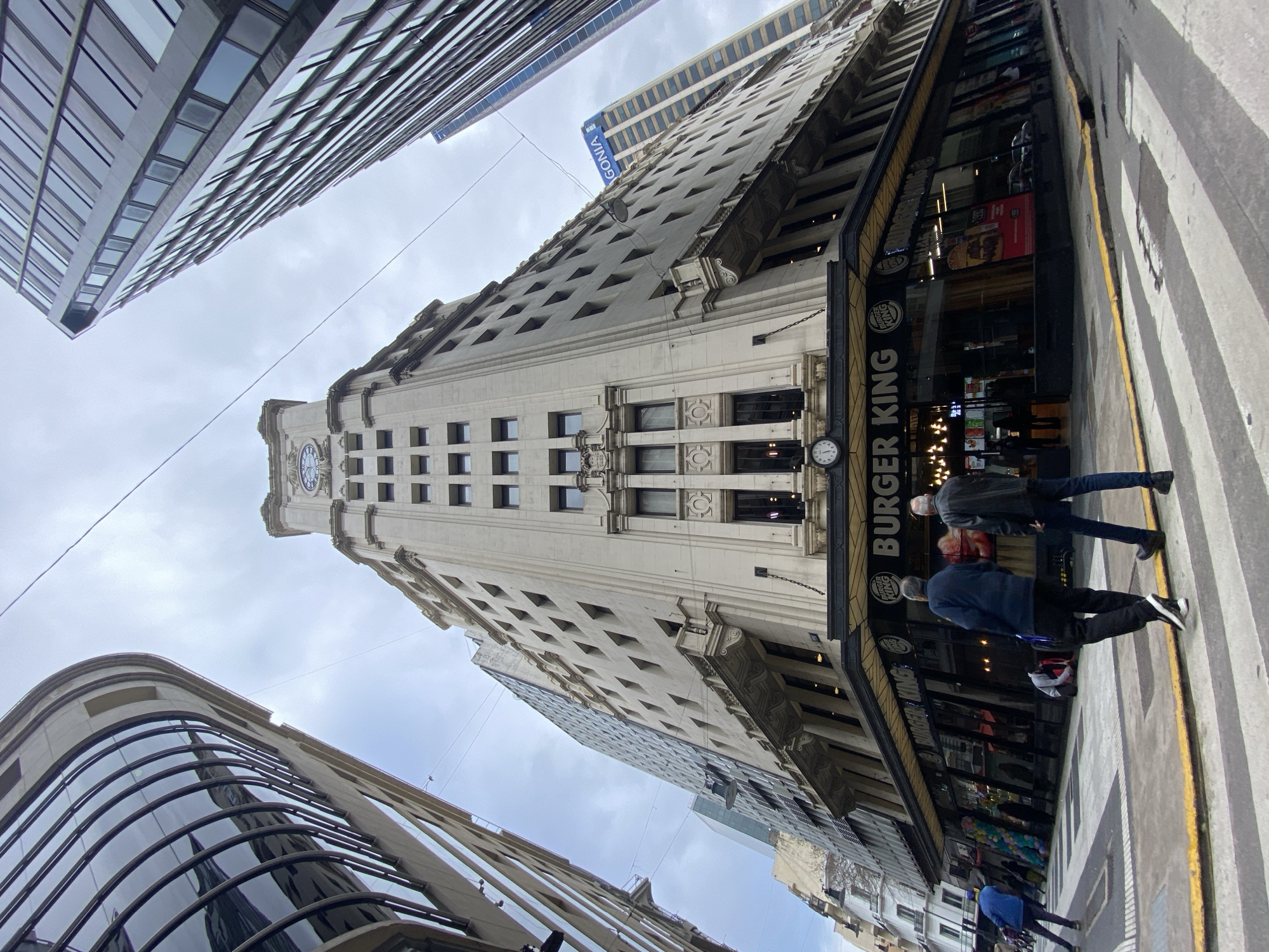 This is a low-angle, wide-angle photograph of a Burger King restaurant located on the ground floor of a large, ornate, cream-colored building with a clock tower at the top. The building is situated in Buenos Aires, Argentina, likely in a downtown commercial district, as evidenced by the surrounding modern skyscrapers and the urban streetscape. The Burger King's facade is predominantly glass, displaying advertisements and the restaurant's interior.
Two men are walking across a crosswalk in the foreground, facing away from the camera. They are dressed casually, one in a dark jacket and jeans, the other in a dark blue jacket and dark pants. Another person in a bright blue jacket is rollerblading on the left side of the frame, further down the street. In the middle ground, near the entrance of the Burger King, a person is kneeling on the sidewalk. Other pedestrians are visible through the glass facade of the restaurant.
The street in front of the Burger King has white and yellow painted lines for a crosswalk and is paved with asphalt. The sky is overcast, contributing to the muted lighting of the scene. The architecture of the buildings on either side of the Burger King varies, with one on the left featuring a curved, modern glass facade, and another on the right being a tall, sleek skyscraper with many windows. Electrical wires are visible strung across the sky. The overall impression is of a busy urban environment.