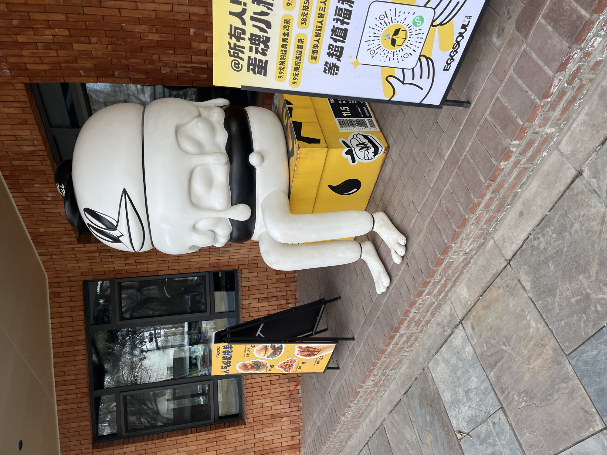 The image captures a street scene in Changchun City, China, featuring a peculiar, cartoonish statue of a figure with an oversized white head, black hair, and white body resting a yellow box. The statue is positioned next to advertisements for a restaurant named "Eggsoul." The surrounding environment includes a brick building with windows reflecting trees, and a sidewalk made of large stone tiles. The picture shows two advertising boards. One on the right side contains a black and yellow EGGSOUL advertisement promoting food deals, a QR code, and the other on the left featuring an image of an EGGSOUL food menu. The image has been rotated and presents the scene in a slightly tilted view.
