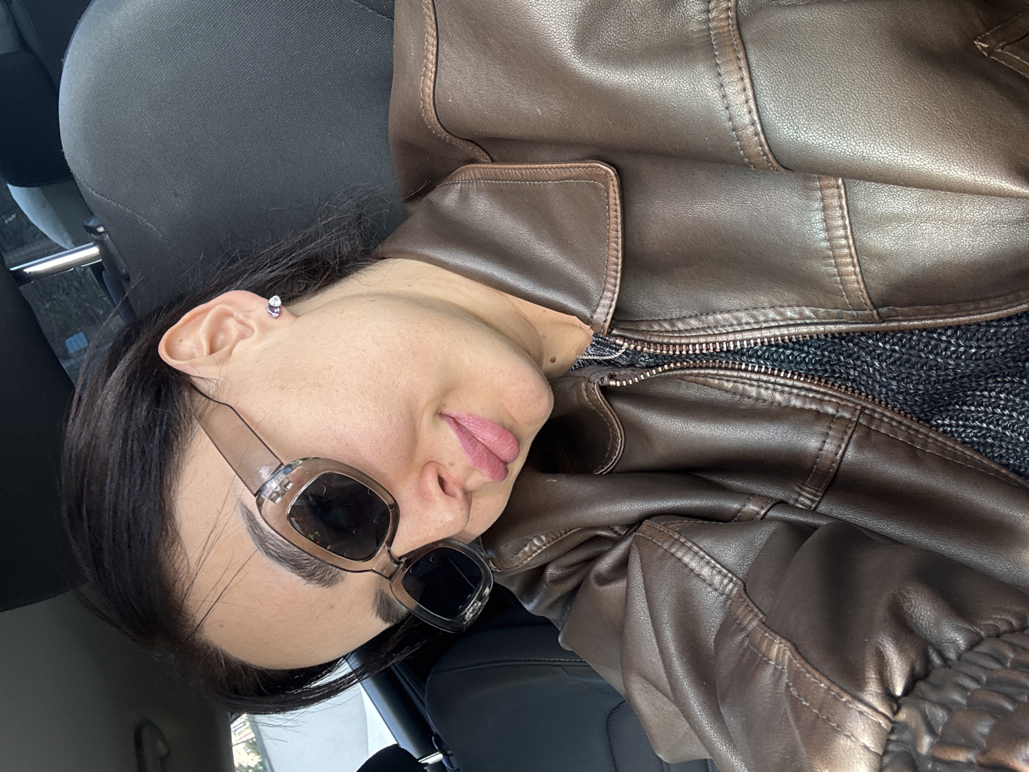 The image is a selfie taken from a low angle of a person inside a car. The person has dark hair, light skin, and is wearing sunglasses. They are wearing a brown leather jacket over what appears to be a dark sweater. The car seat is visible in the background. The location of the photo is unknown, but the prompt specifies Tbilisi, Georgia.