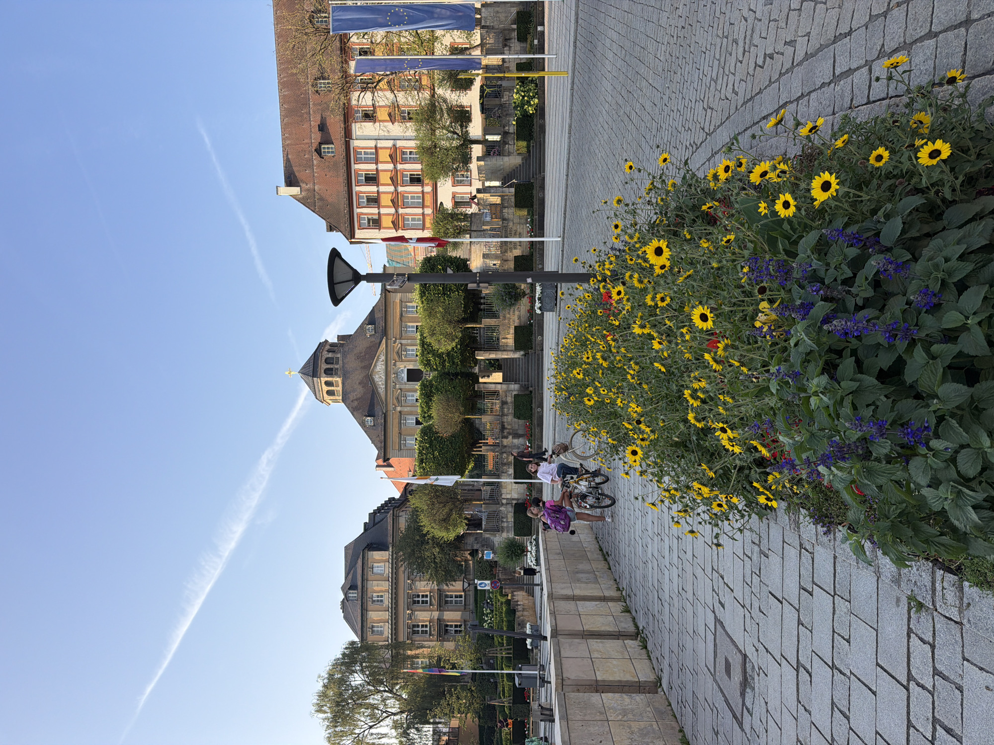The image depicts a street scene in Bayreuth, Germany, likely near a government building or public square, given the presence of flags and formal architecture. In the foreground, a paved walkway is bordered by a flowerbed filled with bright yellow sunflowers and purple flowers. Two young girls are visible on bicycles, one in the foreground with a pink backpack, and another slightly further back. They appear to be interacting, though the specifics of their actions are unclear. Behind the flowerbed and walkway, a row of buildings lines the street. The central building is a large, classical structure with a domed roof and a prominent clock tower. To its left and right are other buildings, one with a lighter facade and mansard roof, and another with a more colorful, orange-and-white exterior. Several flags are displayed, including what appears to be the European Union flag and a red and white checkered flag. The sky is clear with a thin contrail stretching across it. The scene is well-lit, suggesting it was taken during daylight hours, likely in the late afternoon due to the angle of the sun.