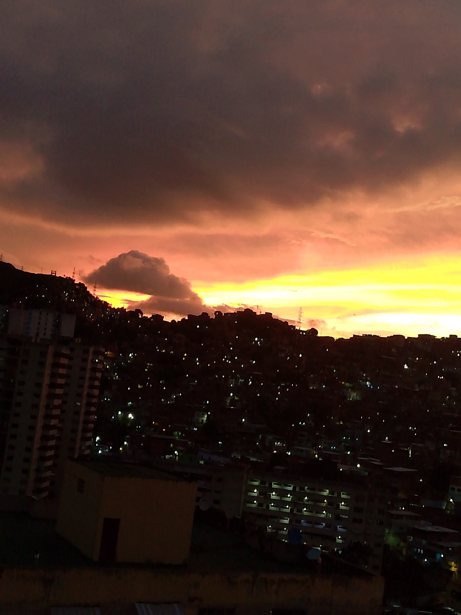 This image captures a twilight scene over a densely populated urban hillside in Caracas, Venezuela. The sky is dominated by dramatic clouds, illuminated with hues of orange and yellow from the setting sun, contrasting with dark, ominous clouds above. Below, the hillside is a sprawling tapestry of buildings, many appearing to be informal settlements, densely packed together. The lights of the city are beginning to twinkle, indicating the transition from day to night.
In the foreground, a few distinct structures are visible. On the left, a tall, modern apartment building stands out, its windows dark or reflecting the dim light. To the right of this building, a smaller, yellow, single-story structure with a dark doorway is prominent. Further down the hillside, more apartment buildings and a multitude of smaller houses and structures are visible, their lights creating a scattered pattern across the dark landscape. The overall impression is one of a vibrant, yet potentially precarious, urban environment set against a striking natural sky. No people or specific actions are discernible in the image.