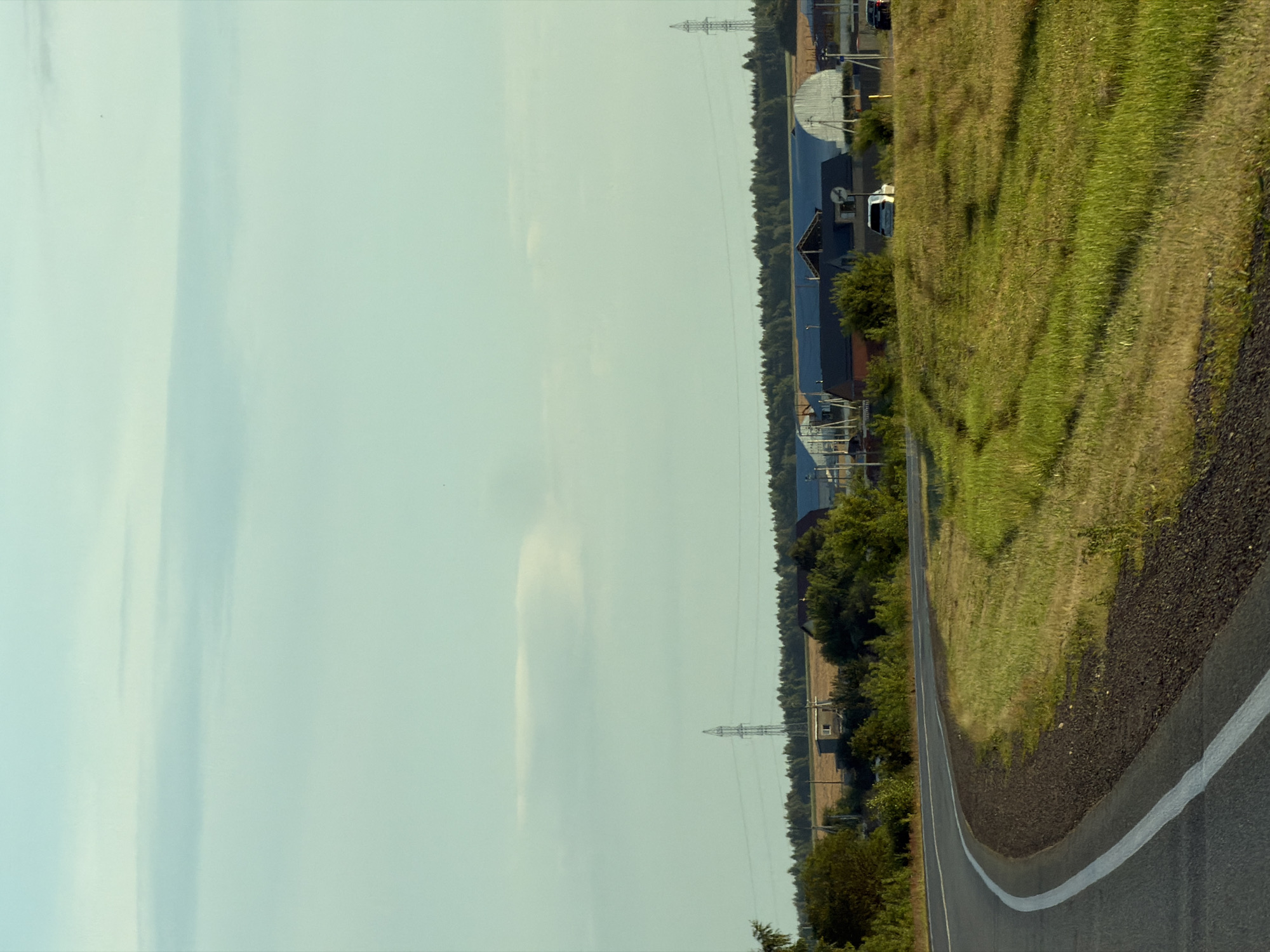 The image depicts a scenic rural landscape near Togashevo, Russia, under a pale blue sky with wispy clouds. The foreground is dominated by a curved asphalt road with white lane markings, transitioning into a grassy embankment. In the midground, a line of trees separates the road from a cluster of buildings, including what appear to be agricultural structures with white, arched roofs and other residential or utility buildings. Power lines and transmission towers are visible in the distance, suggesting the presence of infrastructure in the area. A few vehicles are present, including a white SUV and a dark-colored car, suggesting light traffic. The overall impression is of a peaceful, functional rural setting.