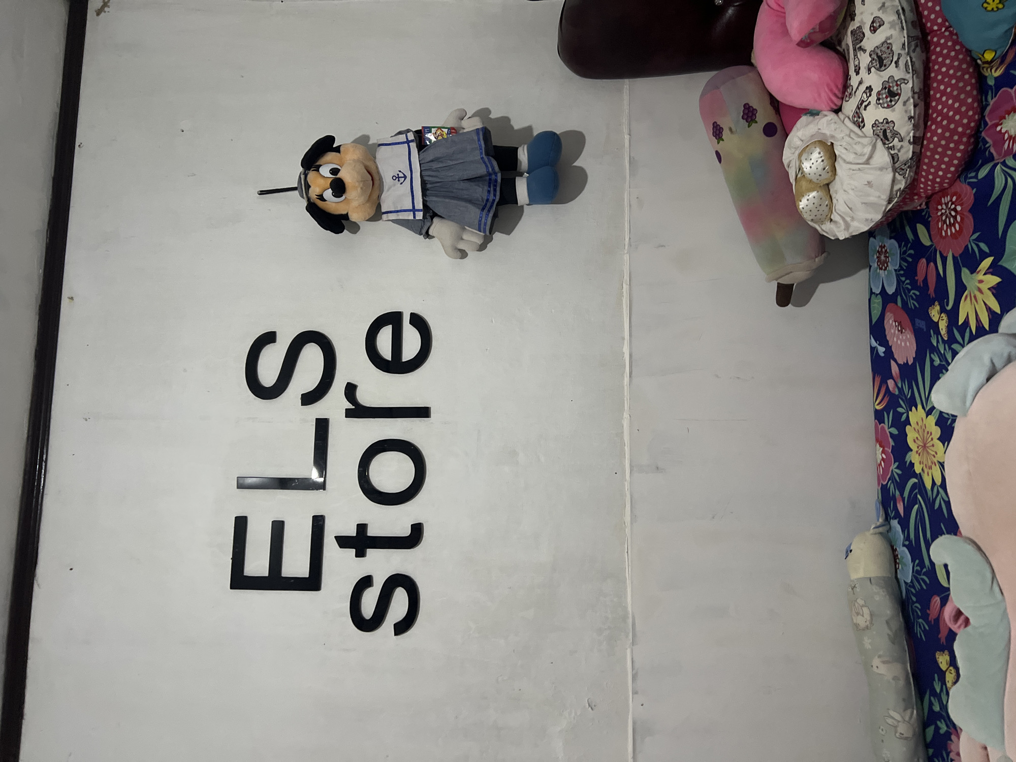 The image shows a wall in what appears to be a domestic setting, possibly in Kecamatan Pati, Indonesia. On the wall, the words "ELS store" are displayed in large, black, three-dimensional letters. To the right of the text, a plush toy of Minnie Mouse dressed in a sailor outfit is hanging on the wall. The lower portion of the image shows a bed with colorful floral-patterned bedding. Several pillows are scattered on the bed, including a long cylindrical pillow with a cartoon pattern, and a stack of circular cushions. A portion of a dark brown chair is visible on the far right of the frame. The wall itself is textured and painted white, with a horizontal line marking a transition in the surface texture lower down.