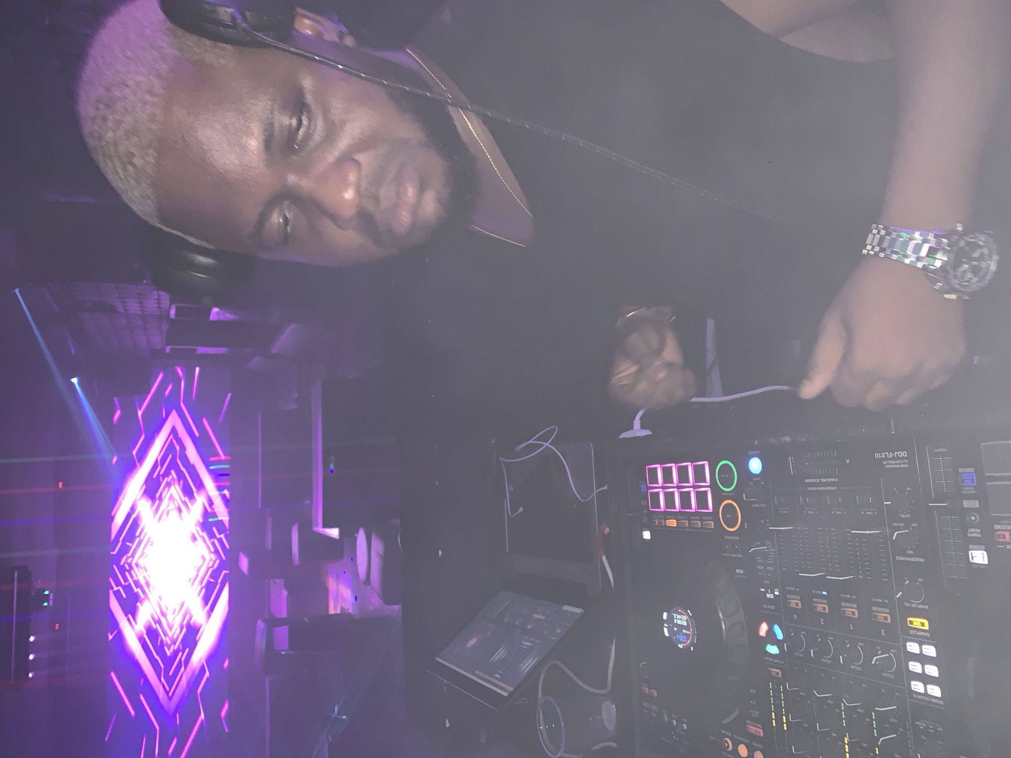 In Howayi, Nigeria, a Black male DJ with bleached blonde hair and headphones on is seen operating a DJ console in a dimly lit nightclub. The DJ is wearing a black shirt and a gold chain. A laptop and a plastic cup are on the console. The background features a large LED screen displaying a geometric, neon purple and pink pattern, with ambient club lighting. The overall scene depicts a nightlife setting.