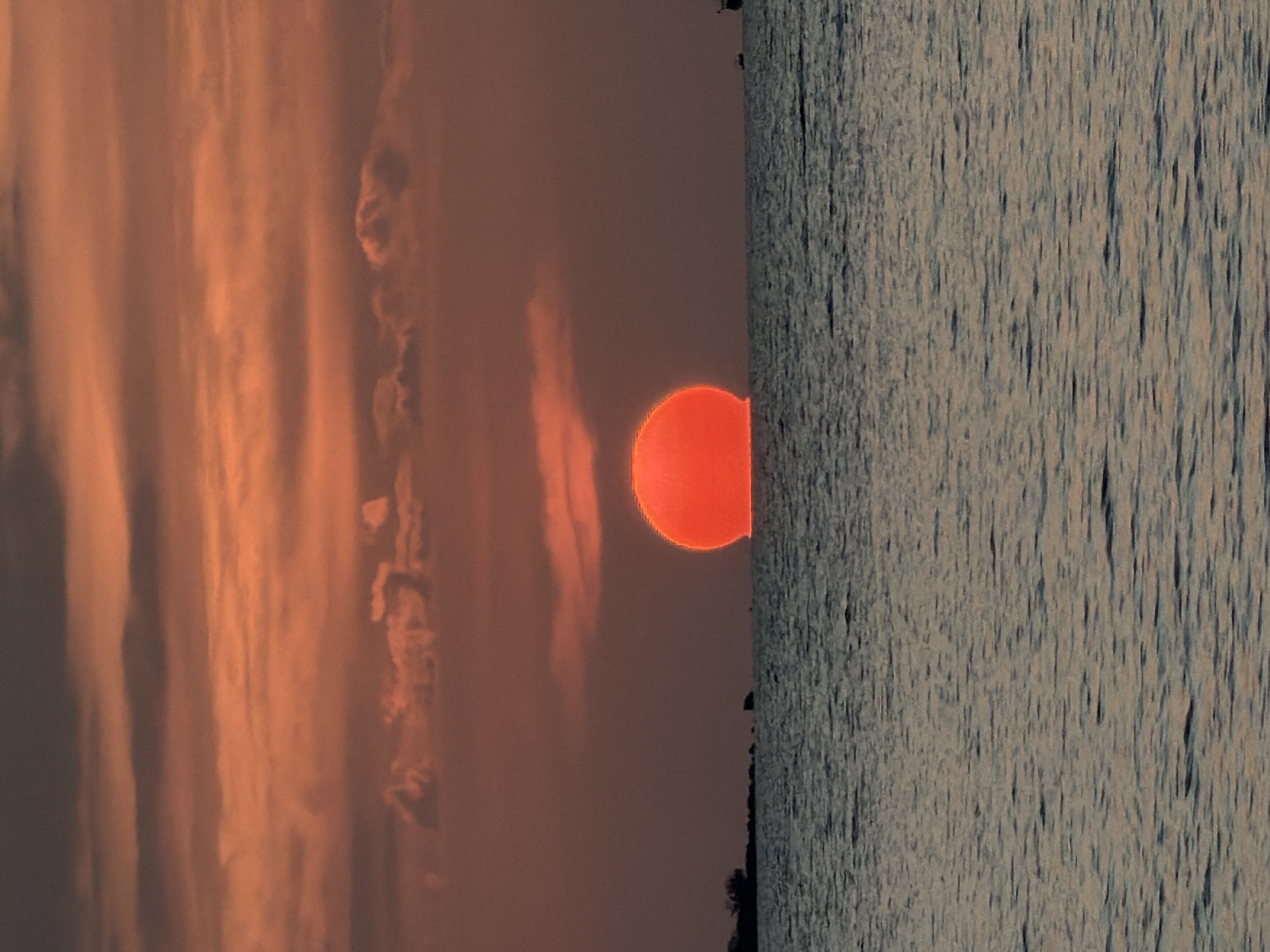 A sunset over the ocean is captured in this vertical, eye-level shot. The sun, a vibrant orange orb, is partially submerged on the horizon, casting a warm glow across the sky. The sky is filled with layers of clouds, tinged with shades of orange, pink, and gray, creating a dramatic backdrop. The ocean below is calm, with gentle ripples reflecting the colors of the sky. In the distance, a dark, silhouetted shoreline with sparse vegetation is visible on the left, while a couple of small boats are subtly present on the right. The overall mood is tranquil and serene, depicting a beautiful natural phenomenon.