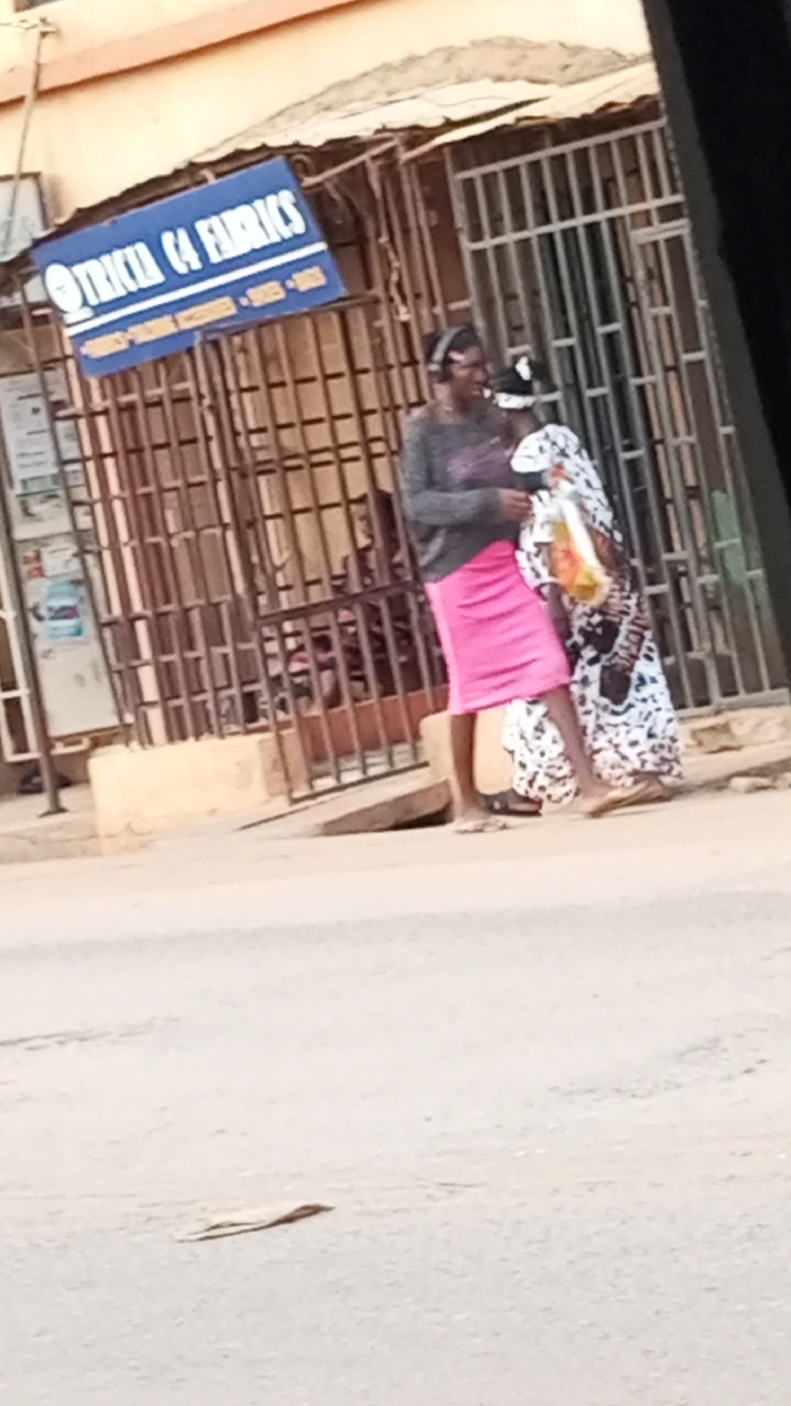The image shows two individuals standing on a sidewalk in front of a business marked by a blue sign reading "TRICIA C4 FABRICS" in Sabon Tasha, Nigeria. One individual is wearing a grey sweater, a pink skirt, and headphones. The other is wearing a patterned white and black dress and carrying a shopping bag. The business has a metal gate. The street is in the foreground.