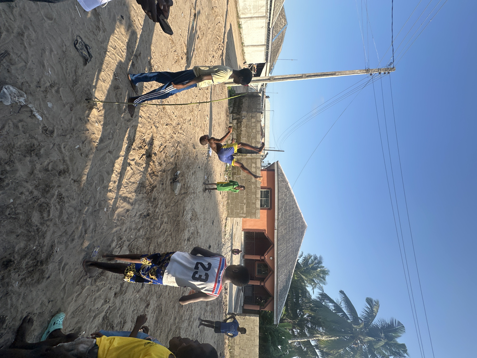 The image depicts a group of young people engaged in a game of limbo on a sandy ground, likely a beach or a dusty street, in Badagry, Nigeria. The sun is high in the sky, casting long shadows, indicating it is likely late afternoon. The weather appears to be clear and sunny.
Several individuals are visible in the frame. In the foreground, a person wearing a white basketball jersey with the number "23" on the back is standing. To the left, another person is partially visible, wearing a yellow t-shirt and blue shorts. In the middle ground, a young boy is in the process of limboing under a thin rope held by two other individuals. He is wearing blue shorts and a yellow shirt. Another child in a green shirt stands further back, observing the activity. The background features a house with an orange-colored exterior and a grey, corrugated roof, surrounded by palm trees. The scene is bordered by brick walls and a utility pole with numerous wires stretching across the clear blue sky.
The subjects appear to be enjoying themselves, with the focus on the energetic limbo game. The setting suggests a warm, casual outdoor environment. The overall mood conveyed is one of youthful exuberance and community activity in a tropical or sub-tropical location.
