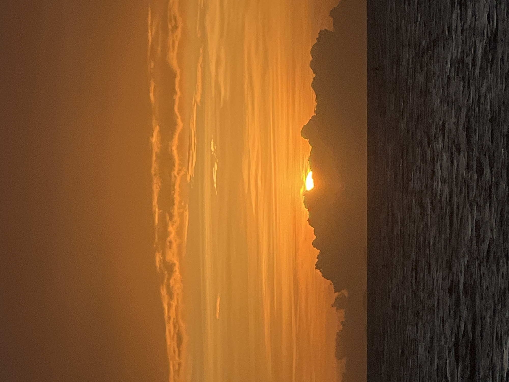 A sunset over the ocean is captured in this vertical, full-frame photograph. The sky is a vibrant gradient of orange and yellow, with streaks of clouds scattered across the upper half of the frame. The sun, partially obscured by a dark, billowing cloud bank, peeks through on the horizon, casting a warm glow. The ocean below is a deep, dark blue, with gentle ripples on its surface. The scene appears tranquil and serene. The location is the coast of Ko Pha-ngan District, Thailand.