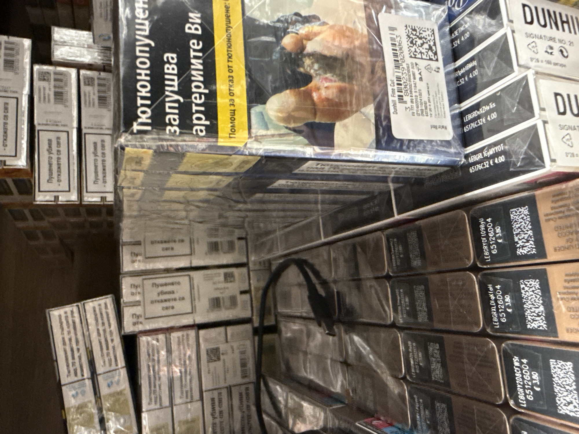 The image shows a collection of cigarette packs stacked on a shelf, likely in a store or storage area. The cigarettes include brands like Dunhill and others with Bulgarian text, implying a possible location in Bulgaria or a market catering to Bulgarian-speaking customers. The cigarette packs display graphic health warnings, including images of diseased body parts and text warning about the dangers of smoking, in Bulgarian. Some packs have pricing information, showing prices in Euros.
