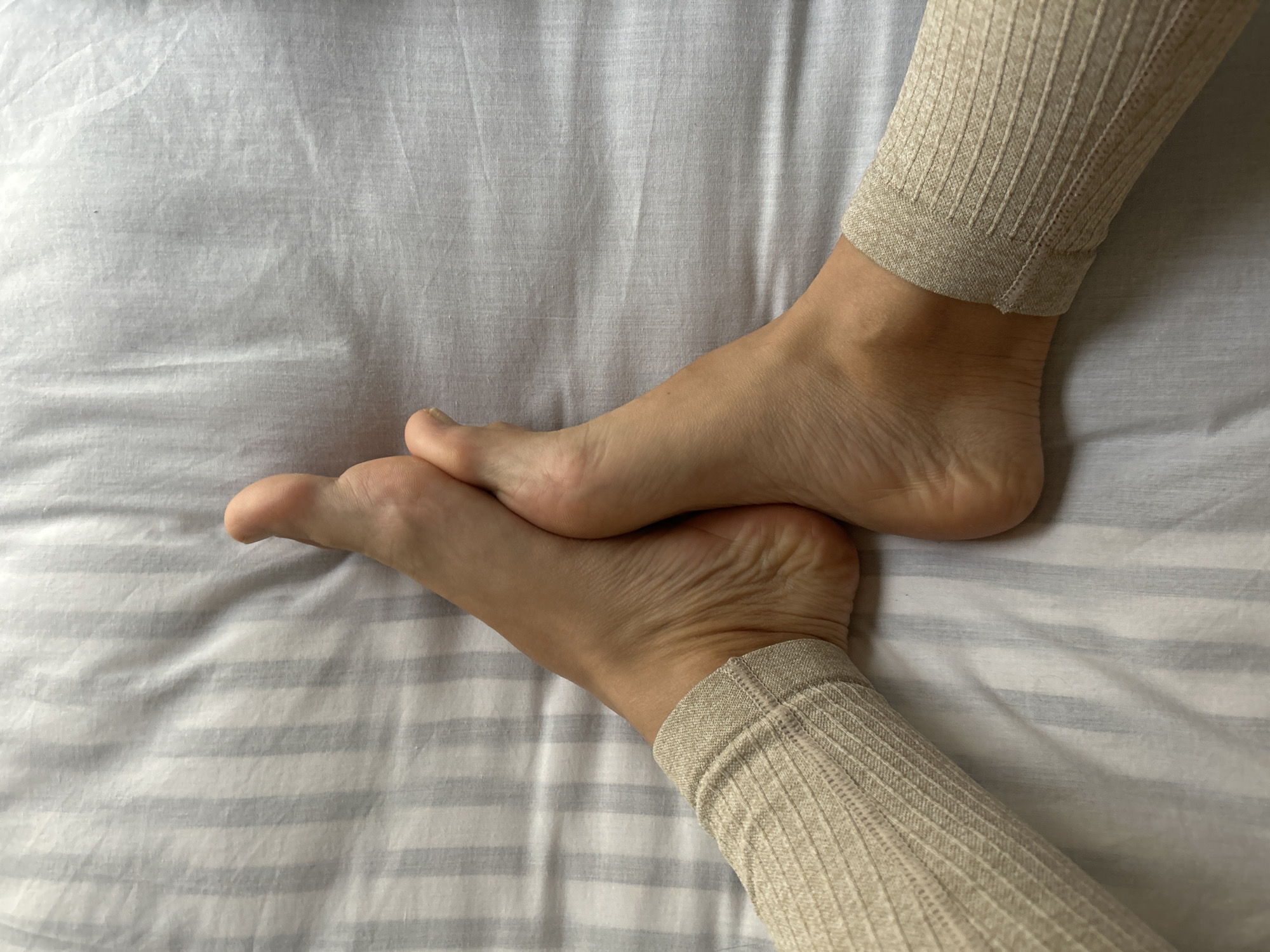 The image shows a pair of feet wearing beige ribbed socks resting on a striped white and grey bedsheet. The feet are crossed at the ankles, with one foot overlapping the other. No location information or city context is discernible from the image. The scene depicts a person in bed.